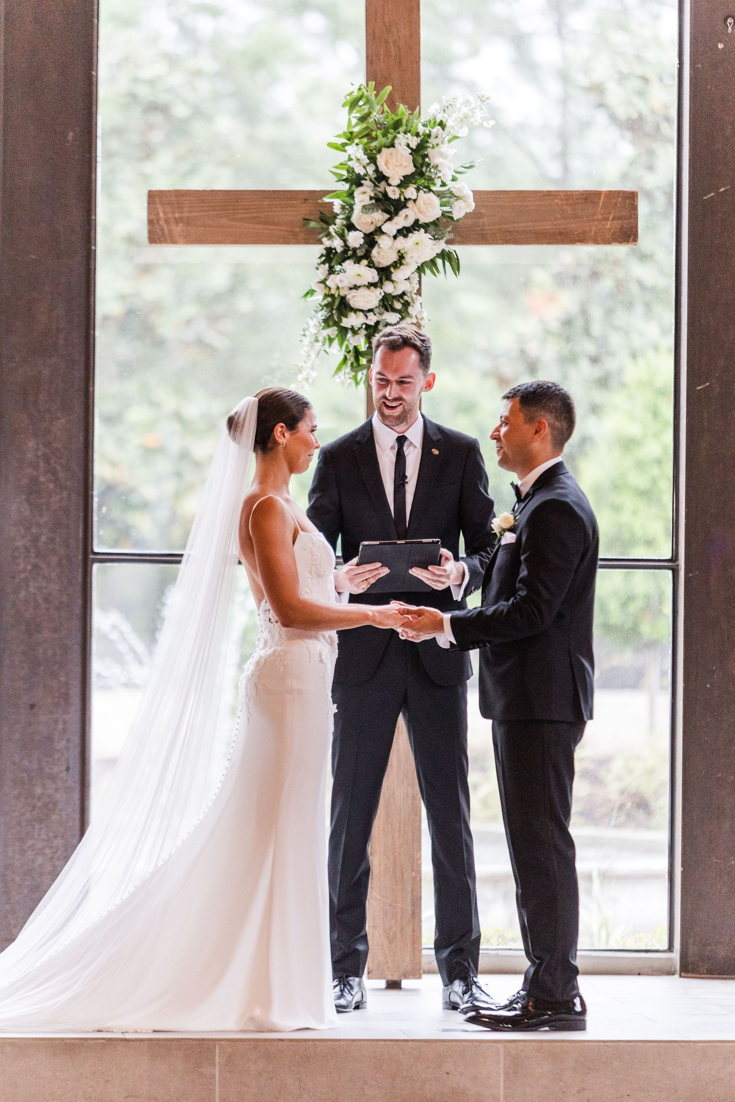 Exchange of wedding vows in front of large windows with elegant floral decorations.