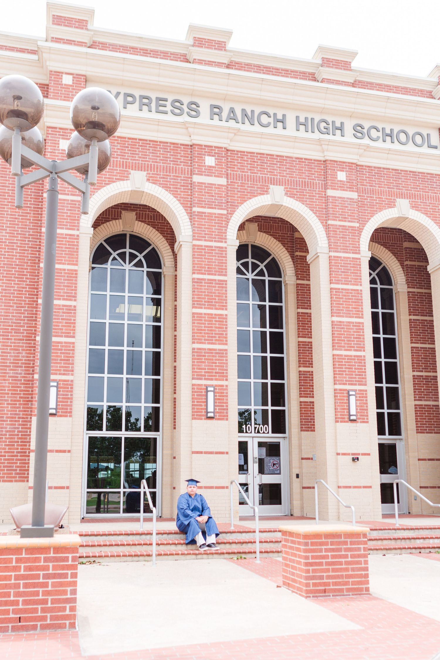 Graduate in cap and gown sits on steps of Cypress Ranch High School brick building.