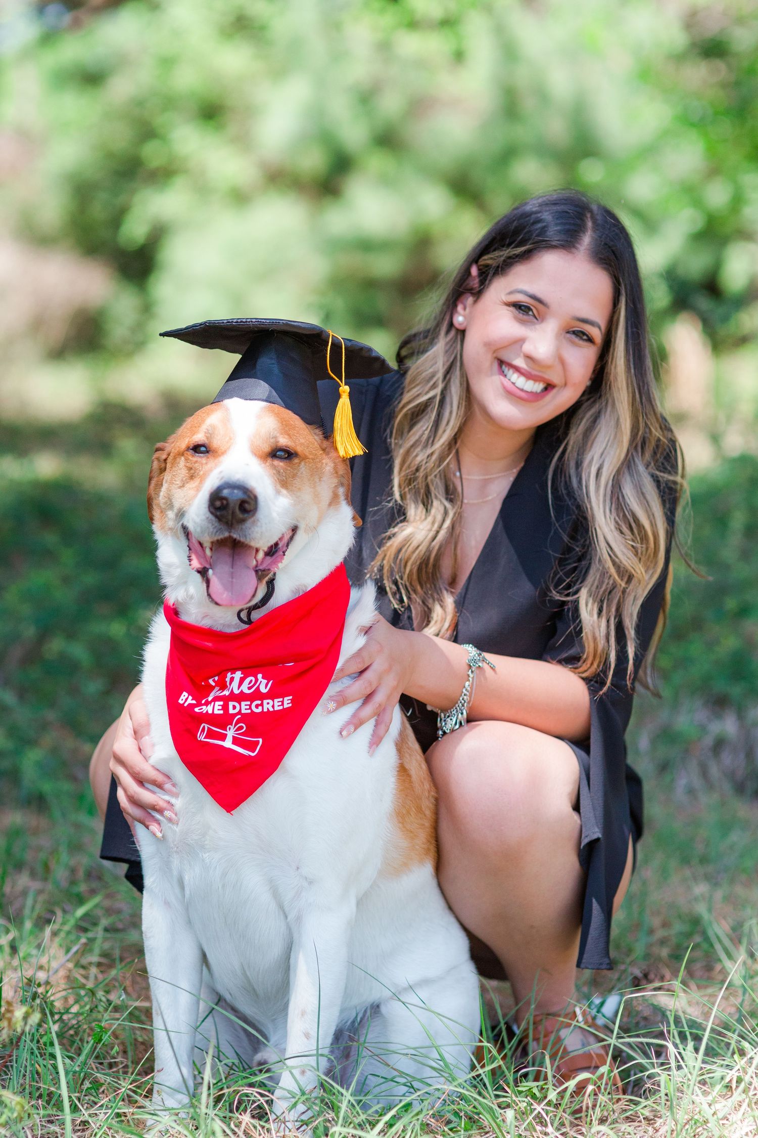 Happy dog wearing graduation cap and red bandana poses with owner in outdoor graduation photo session.