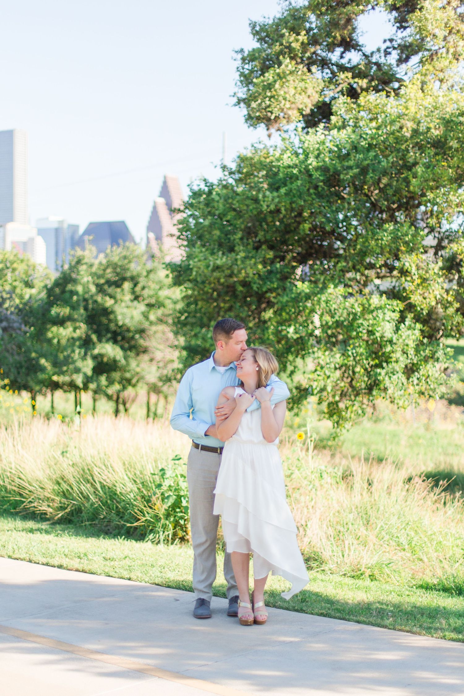 Couple walking along pathway with city skyscrapers visible in background during sunset.