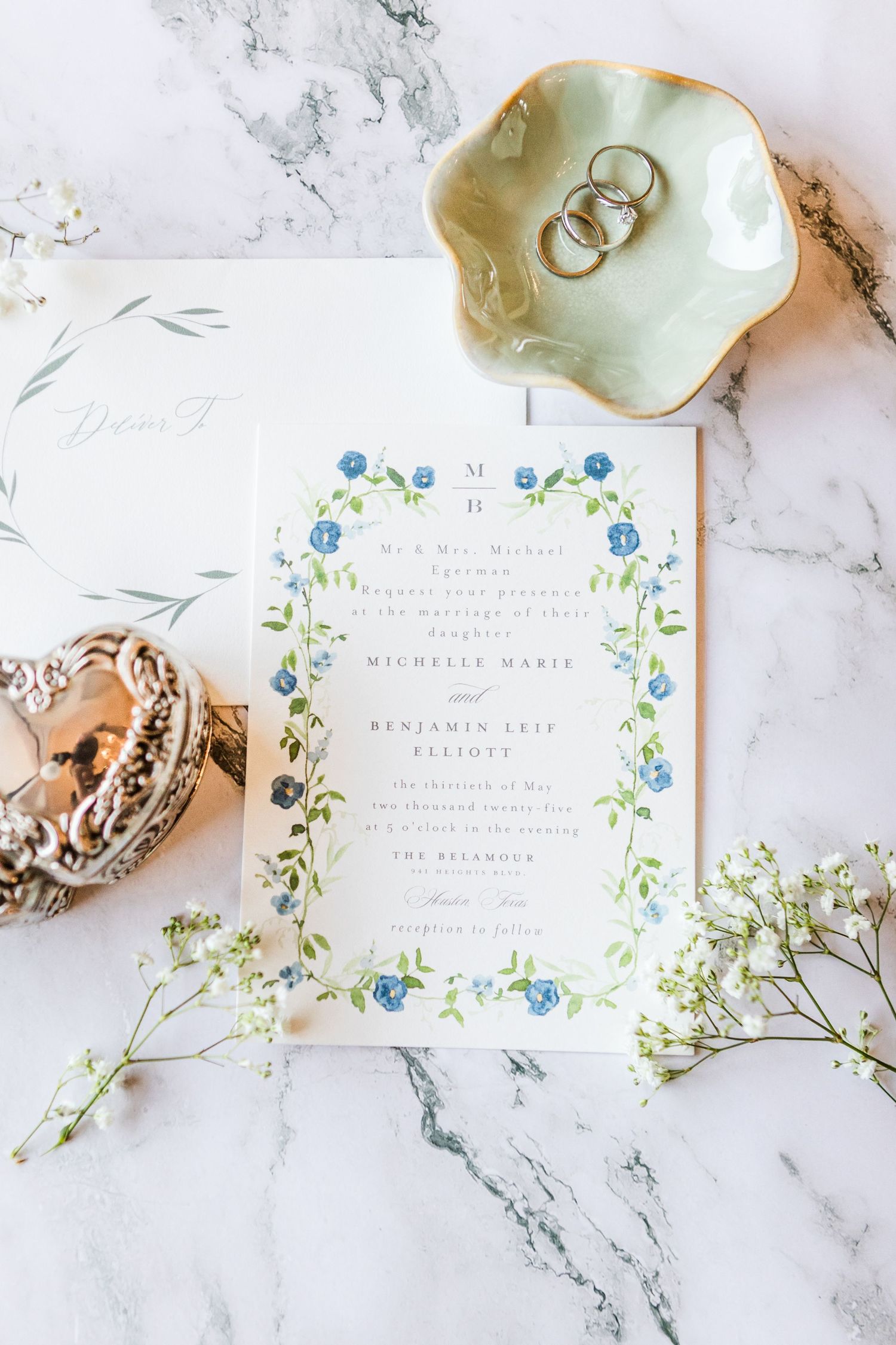 Elegant wedding stationery with blue floral details and rings on marble backdrop.