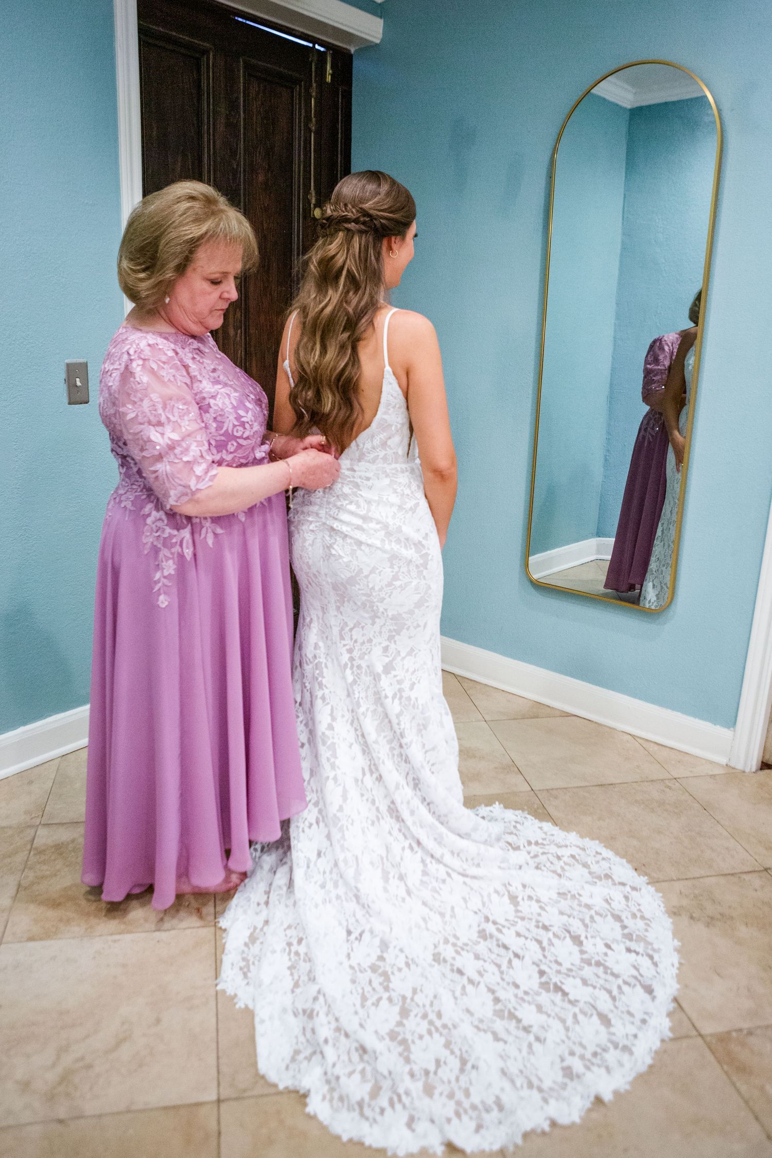 Wedding dress with intricate lace train being prepared in a room with blue walls.