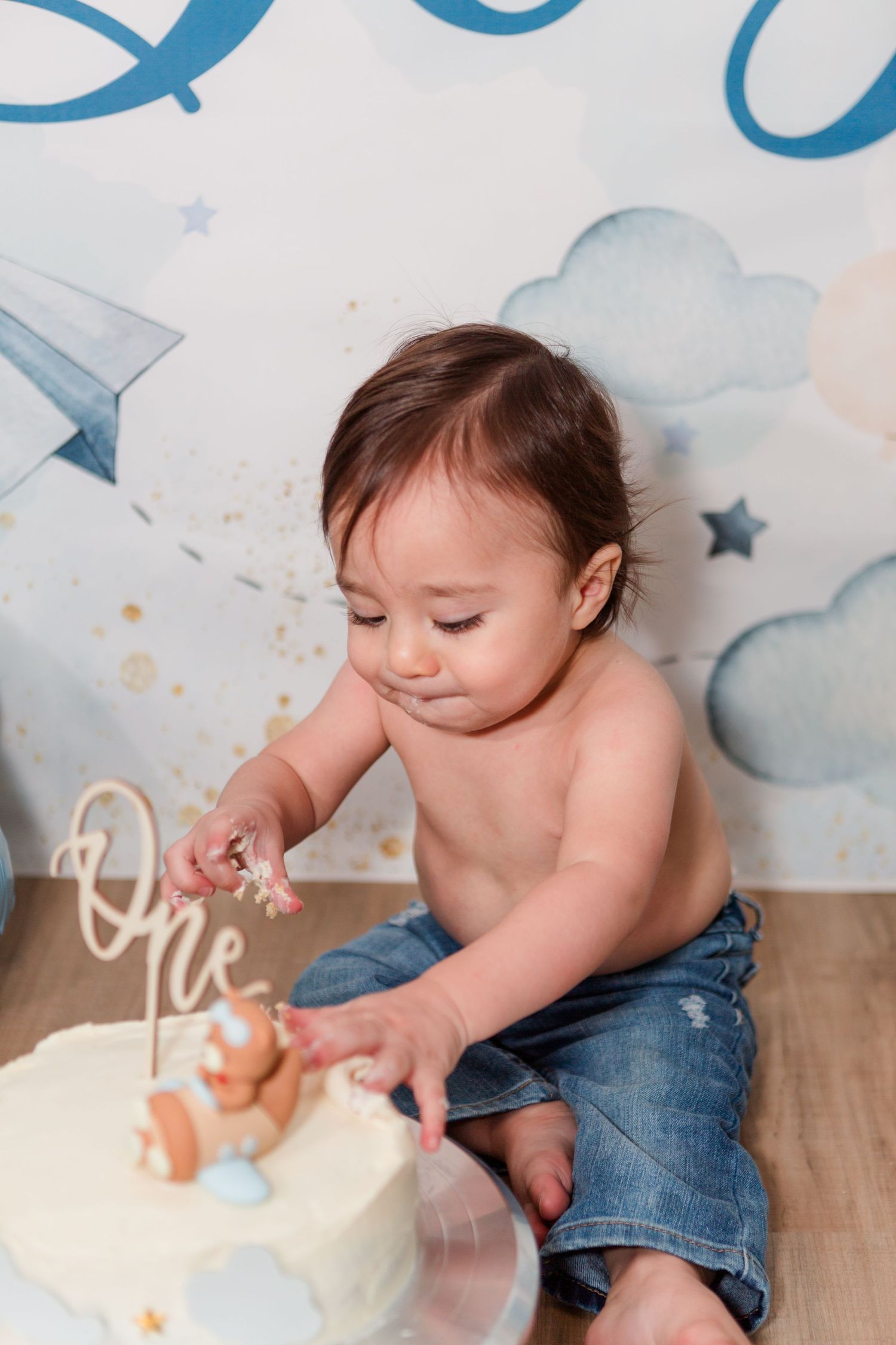 Baby in denim exploring a white birthday cake with blue decor against a cloud-patterned backdrop.