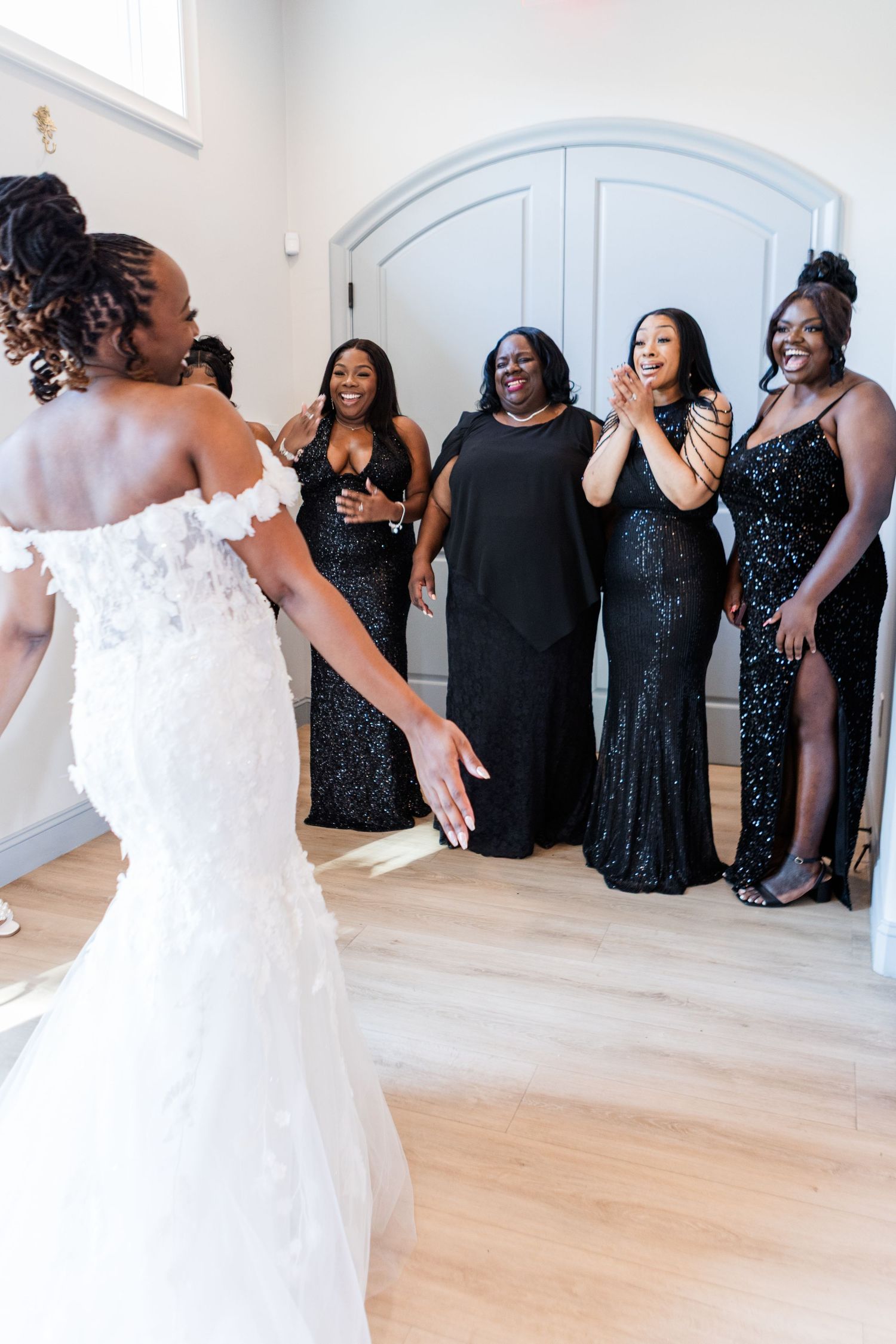 Bridesmaids in sparkly black dresses gathered in a bright white room with hardwood floors.
