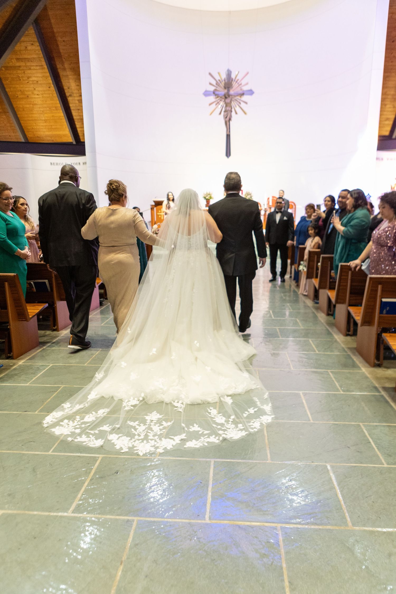 A flowing cathedral length wedding veil trails down the church aisle during a ceremony.
