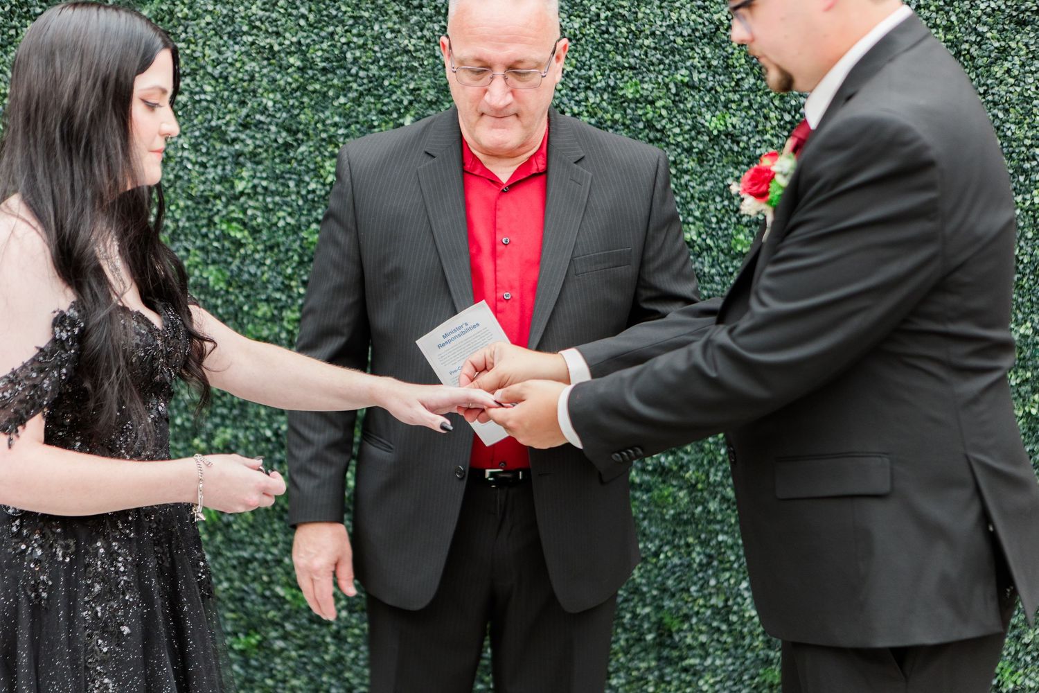 Wedding ceremony taking place in front of green hedge wall with officiant in red tie performing ring exchange.