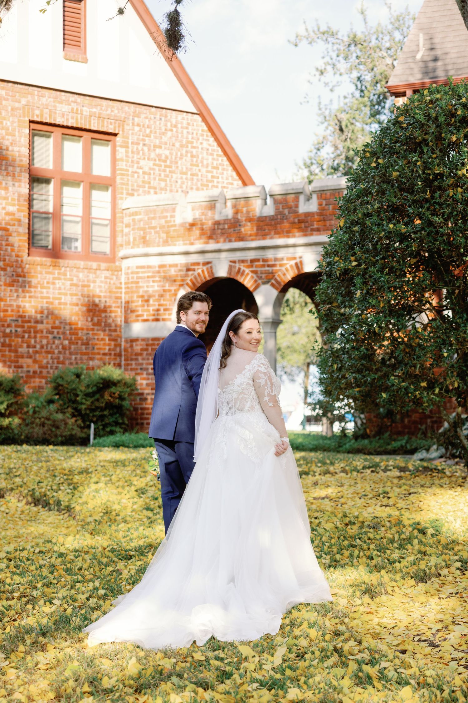 A couple in wedding attire dances and spins on a grassy lawn outside a brick church building in autumn.
