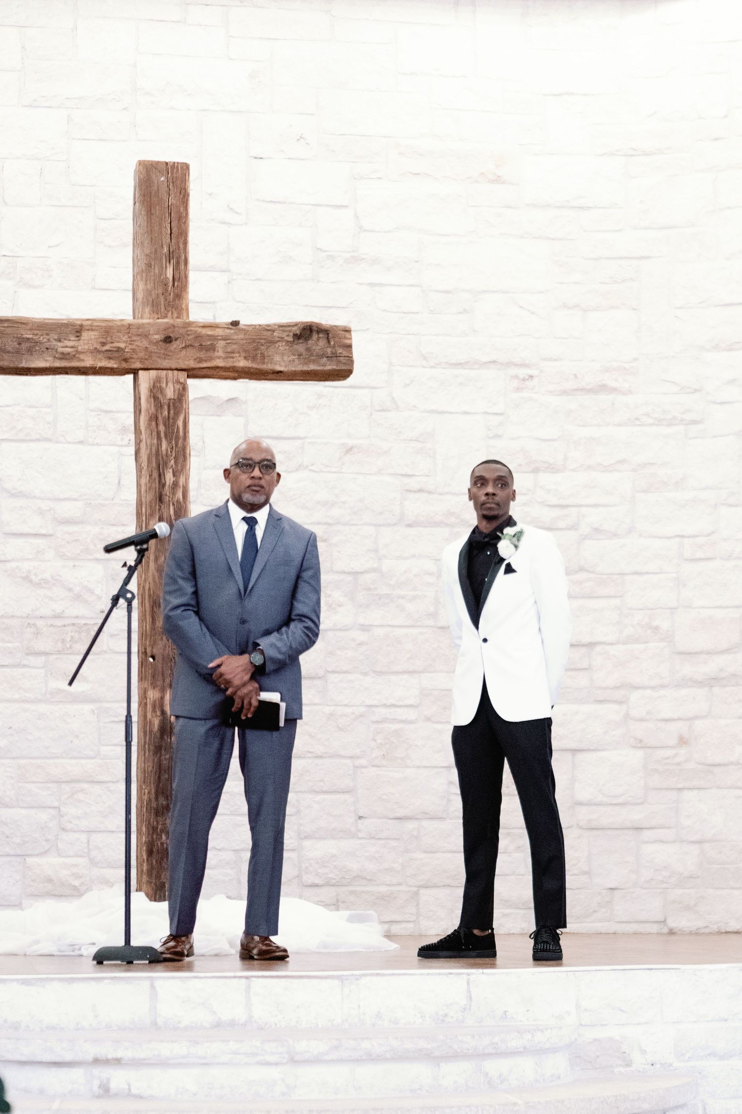 Two people in formal attire stand near a large wooden cross against a white stone wall background.