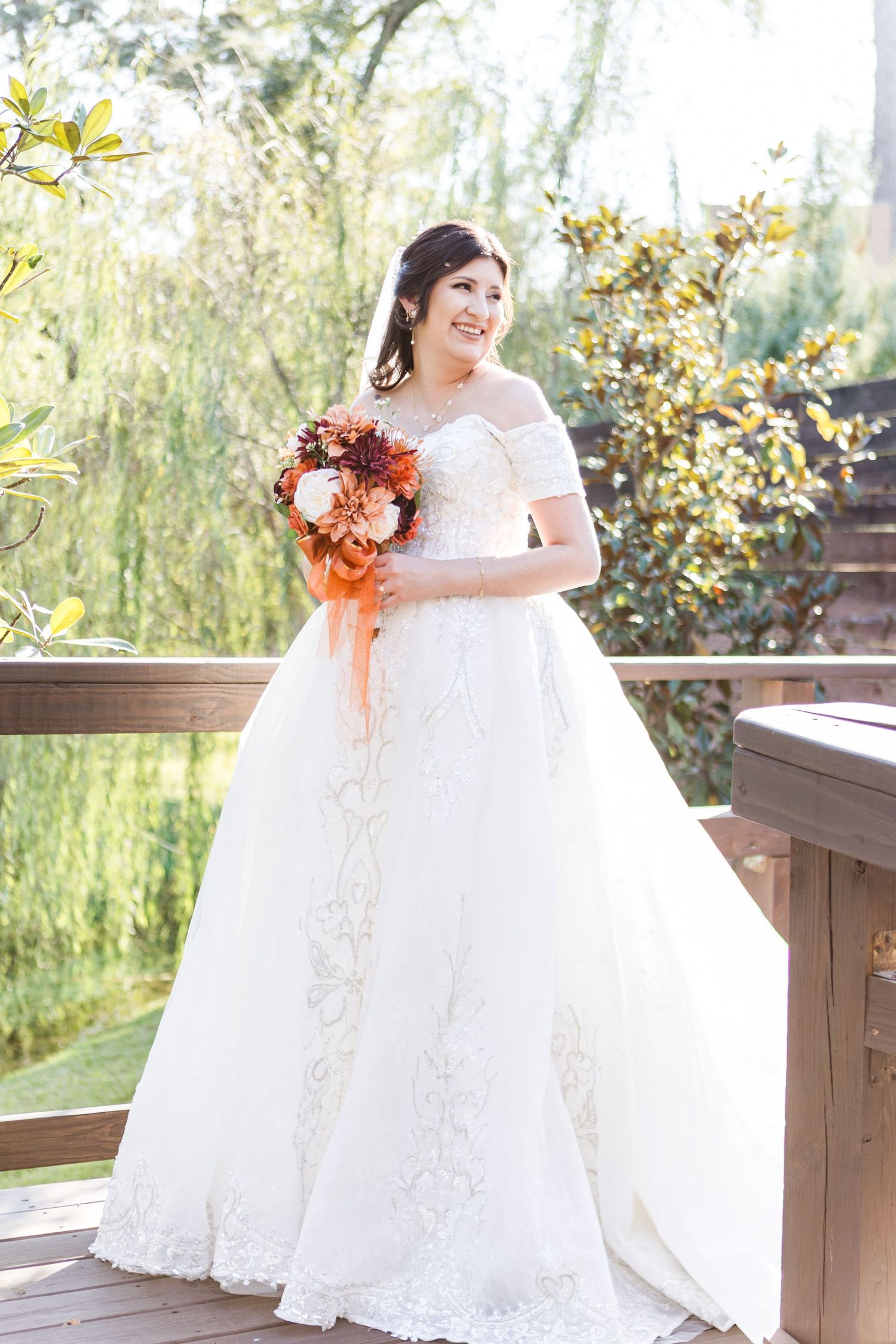A bride in an elegant white ball gown stands on a rustic wooden deck surrounded by autumn foliage.