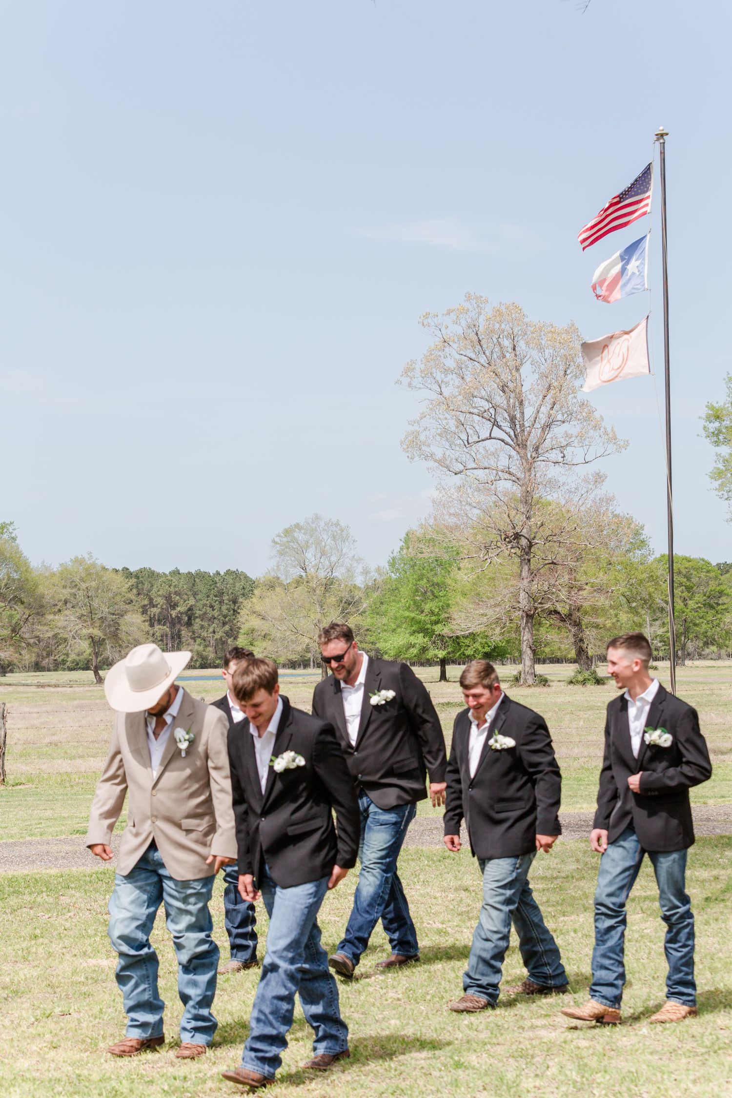 Groomsmen in dark suits and jeans line up in a field on a sunny day.