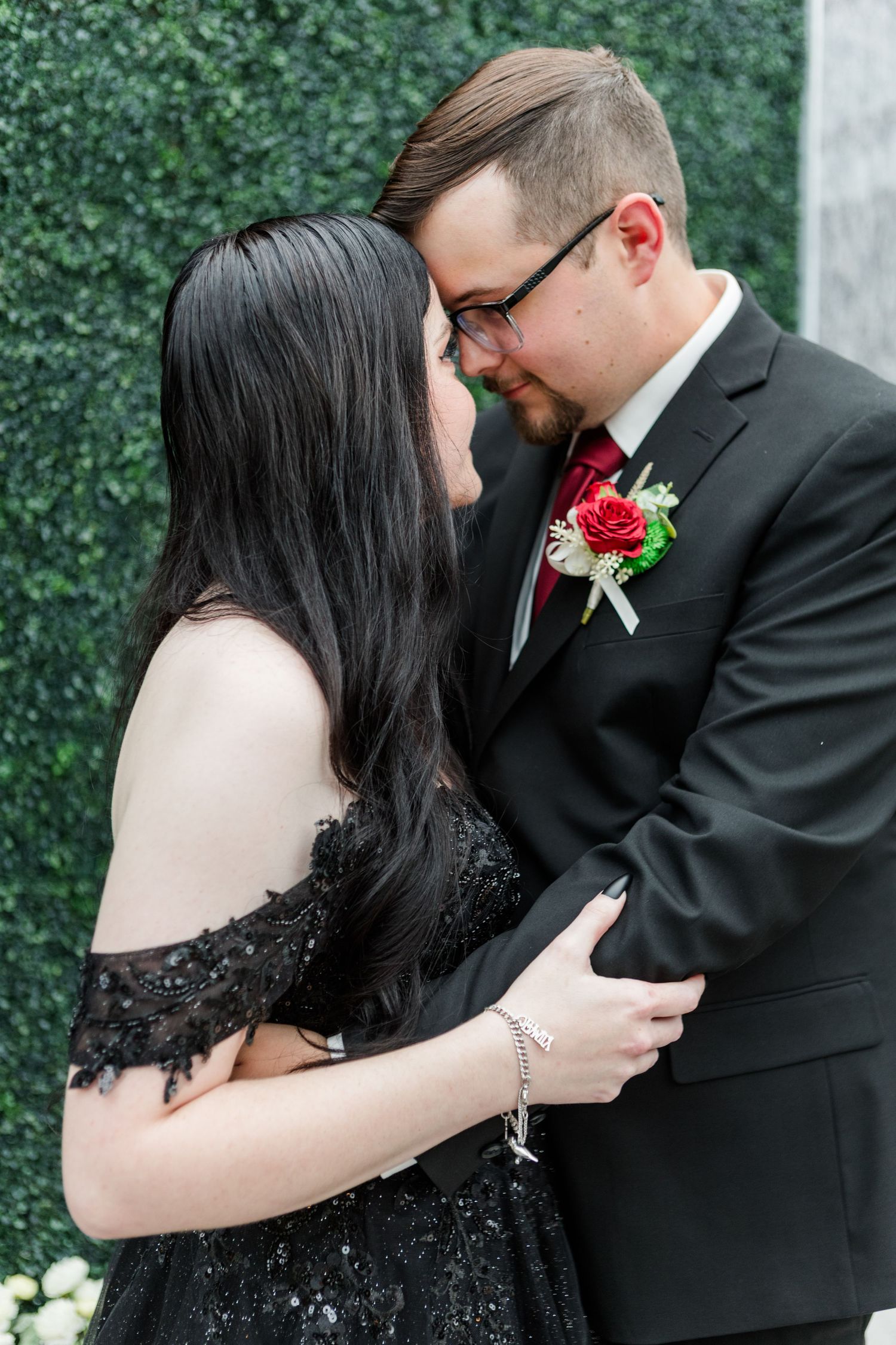Intimate wedding moment captured against green hedge wall with red boutonniere detail.