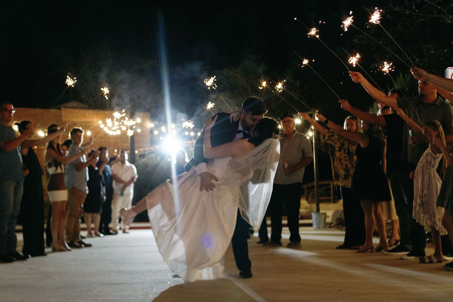 Wedding guests hold sparklers creating a magical nighttime send-off tunnel for the newlyweds.