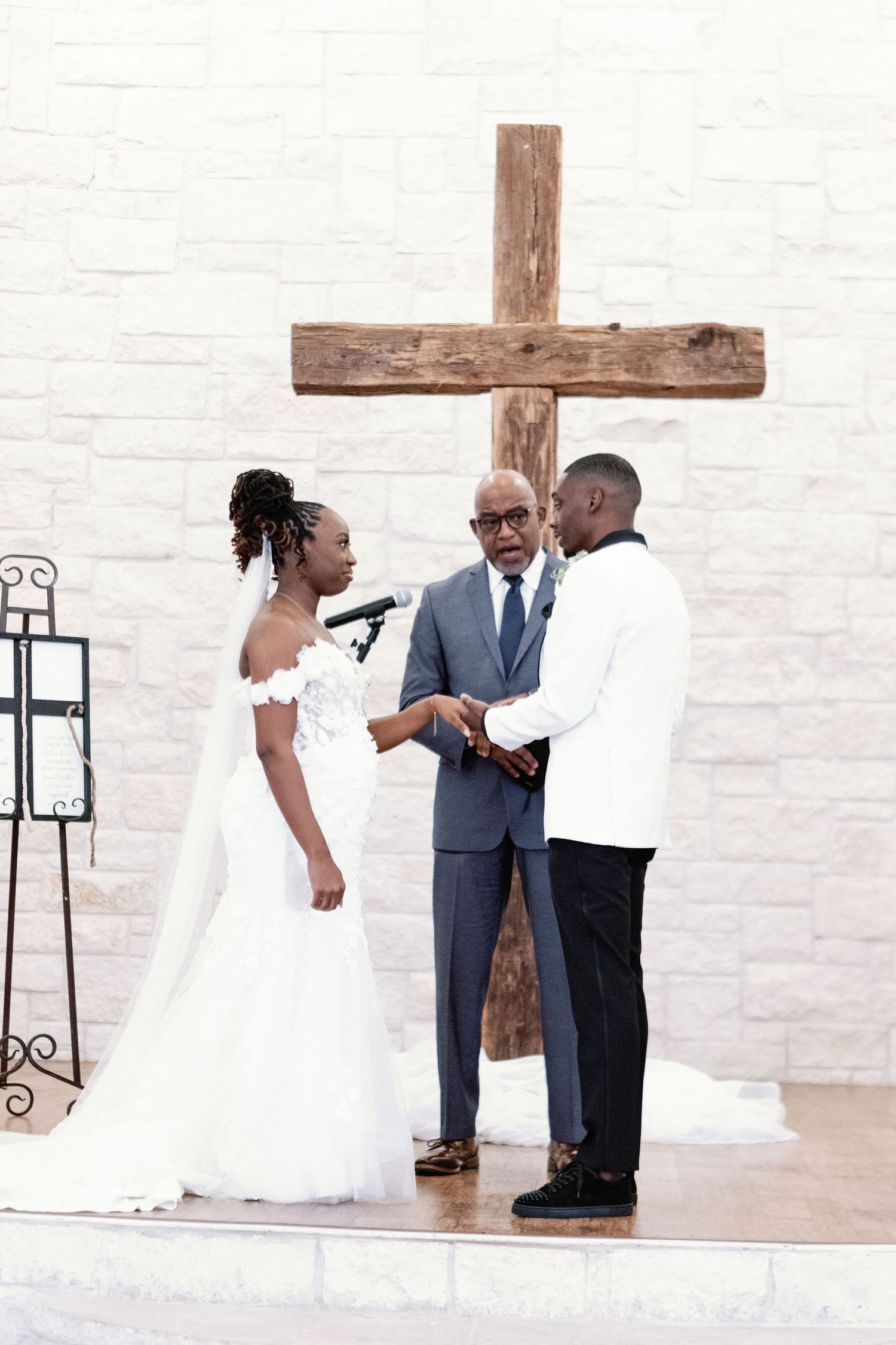 A wedding ceremony scene unfolds in a bright interior space with white walls and a wooden cross mounted behind.