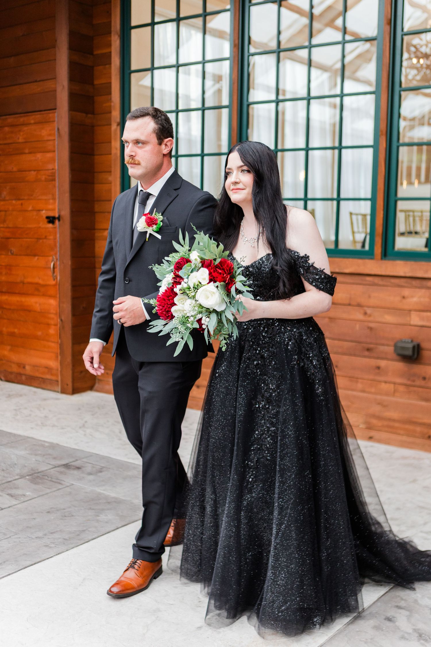 Couple in formal wedding attire standing near wooden exterior and green-trimmed windows.