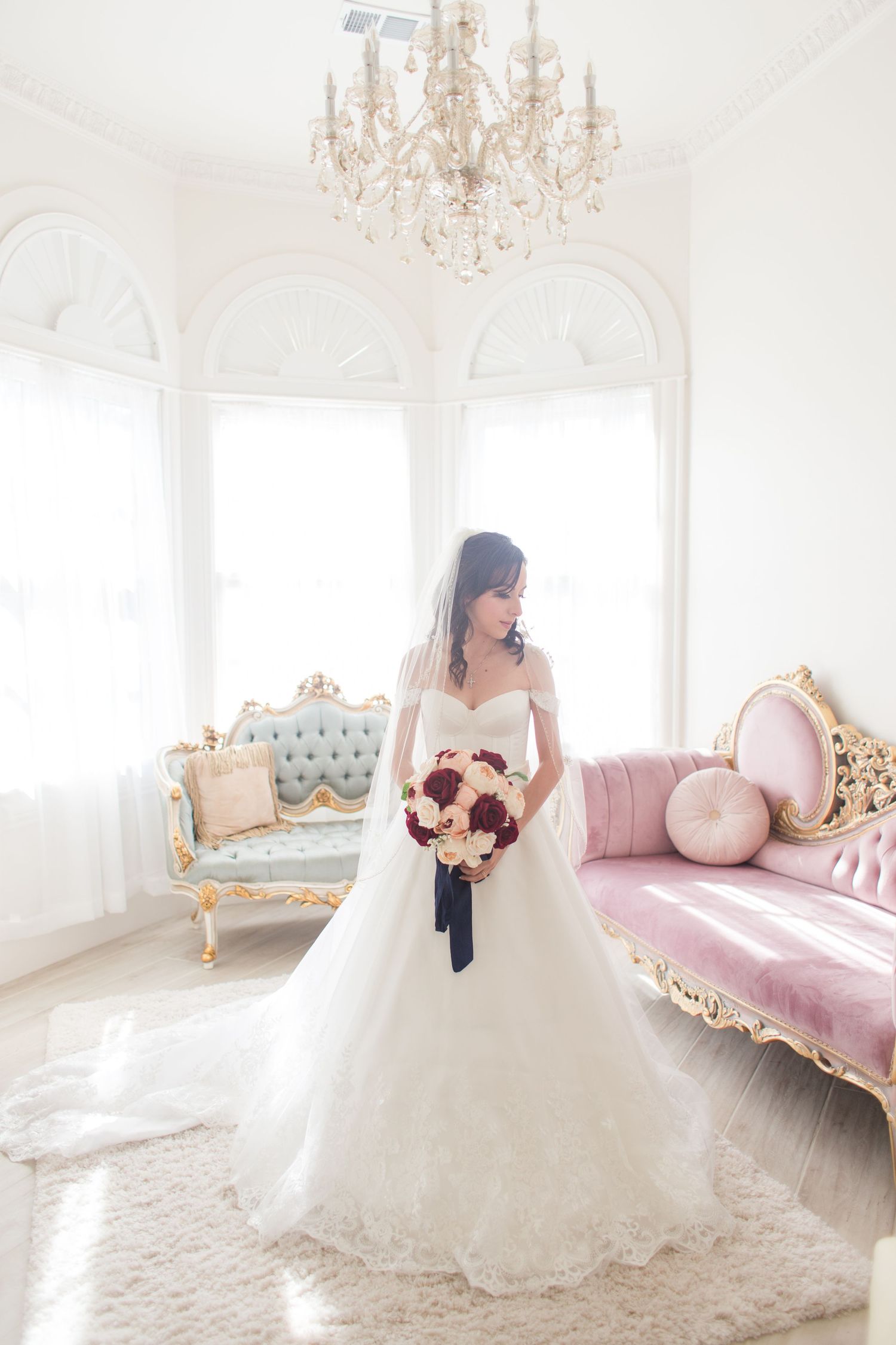 Bride in white ballgown poses on pink vintage chaise in elegant white room with chandeliers.