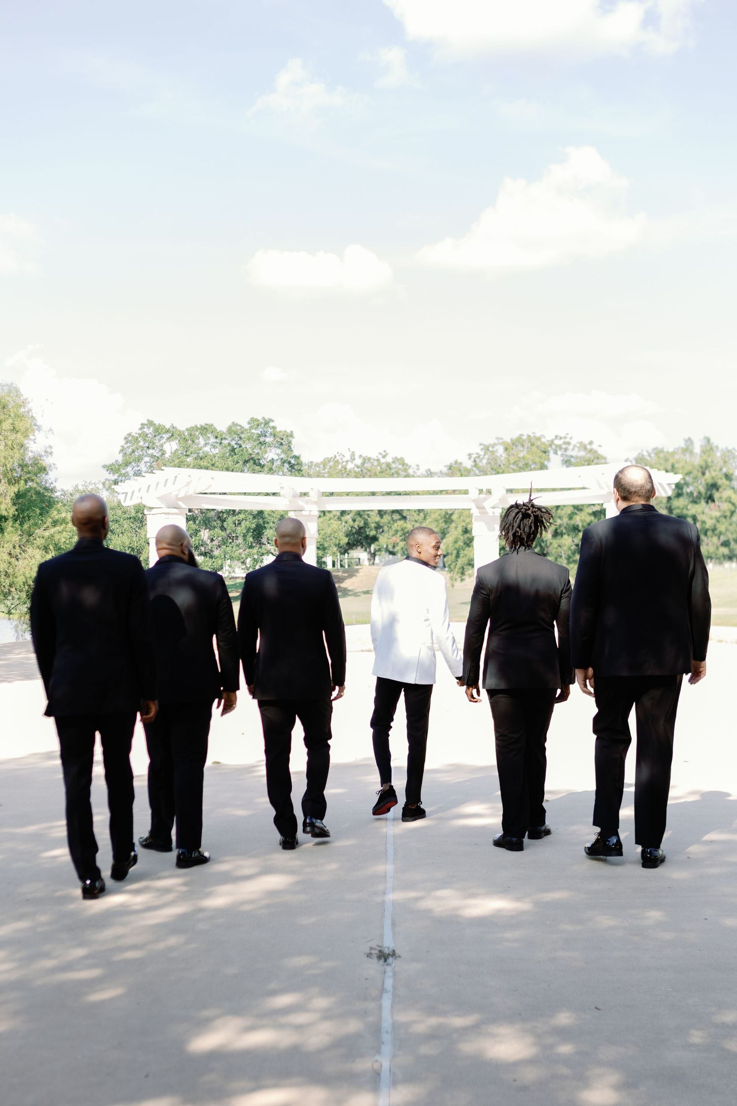 Group of groomsmen in black suits walking together outdoors on sunny day.