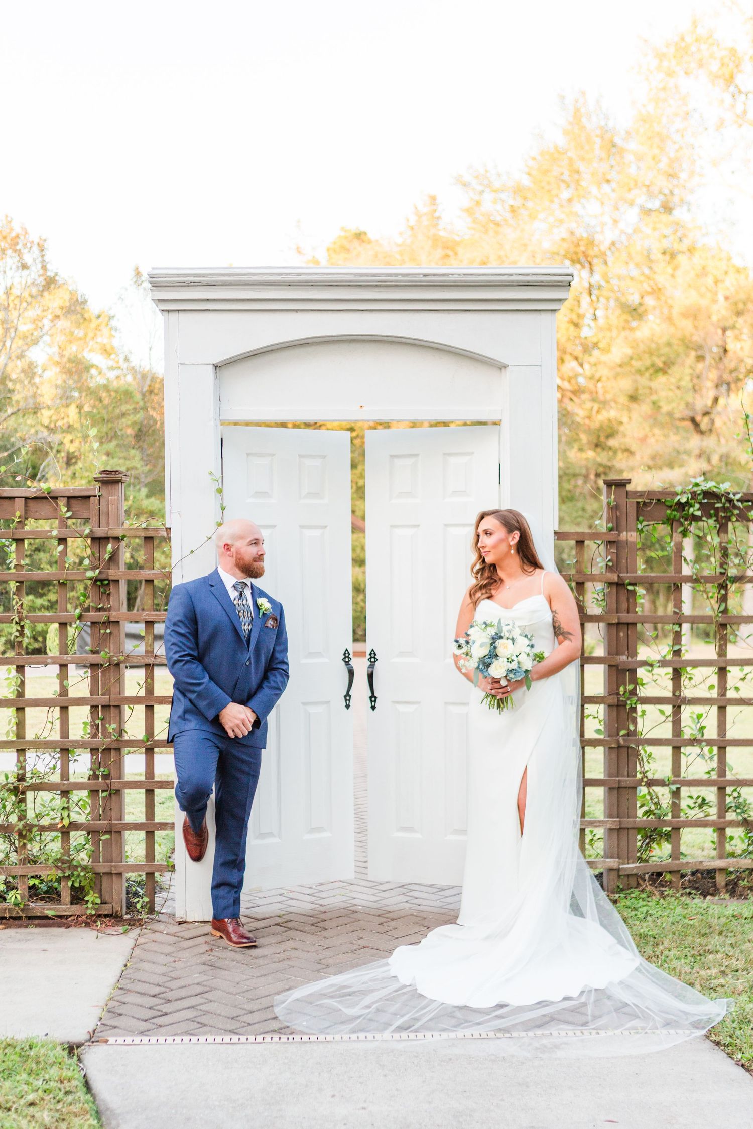Wedding couple poses playfully by white garden doors in outdoor setting.