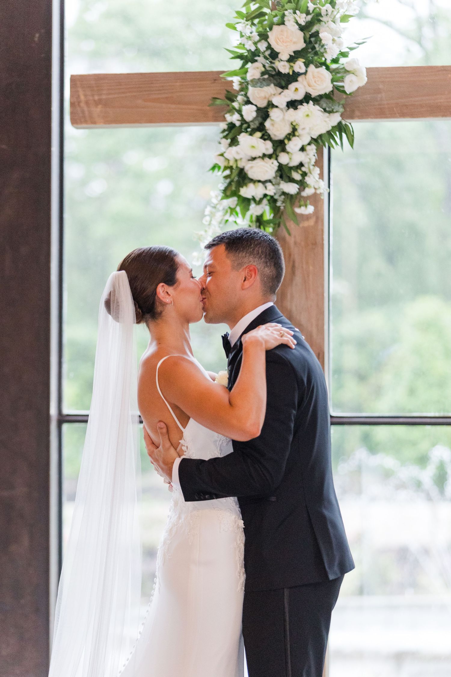 A romantic first kiss as newlyweds during a wedding ceremony with white floral arrangements.