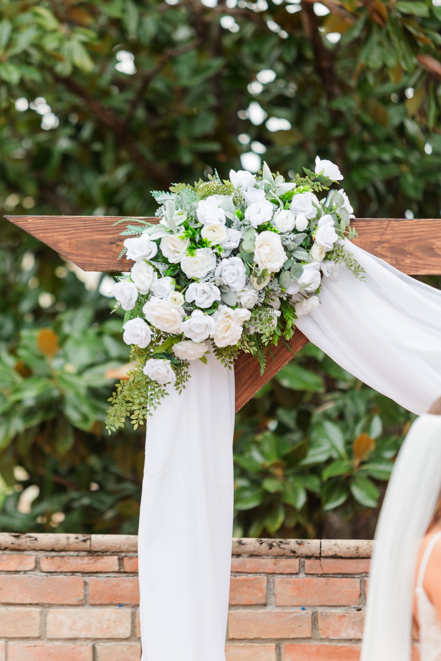 White floral arrangements adorn a wooden arch against a brick wall during an outdoor wedding ceremony.