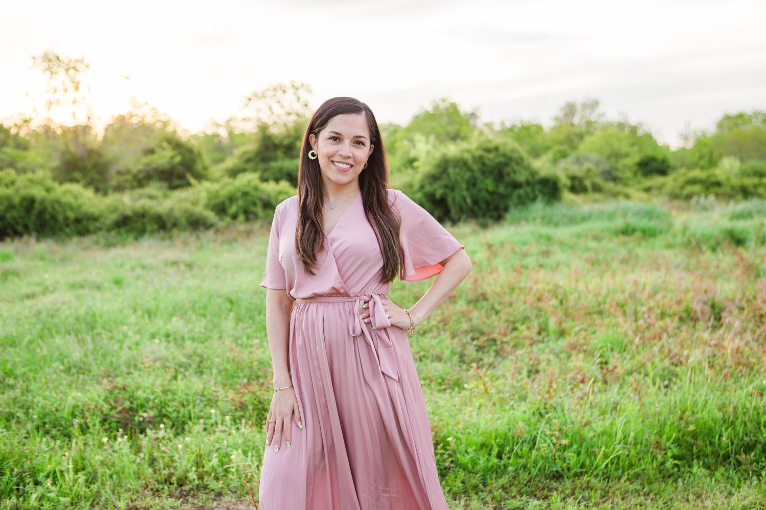 Woman in flowing pink dress posing in a green meadow during golden hour.