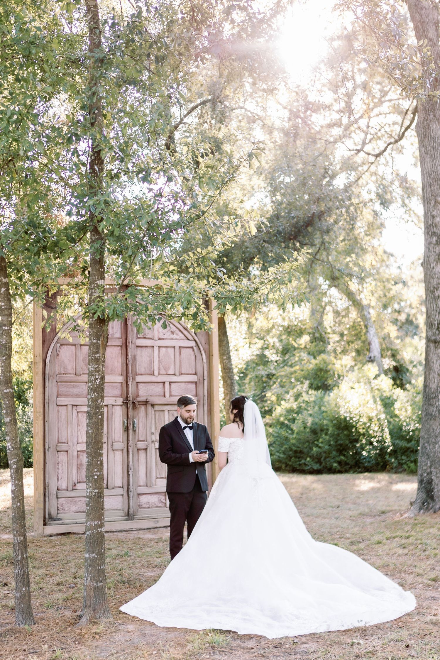 Elegant wedding couple poses in front of vintage wooden doors surrounded by trees.