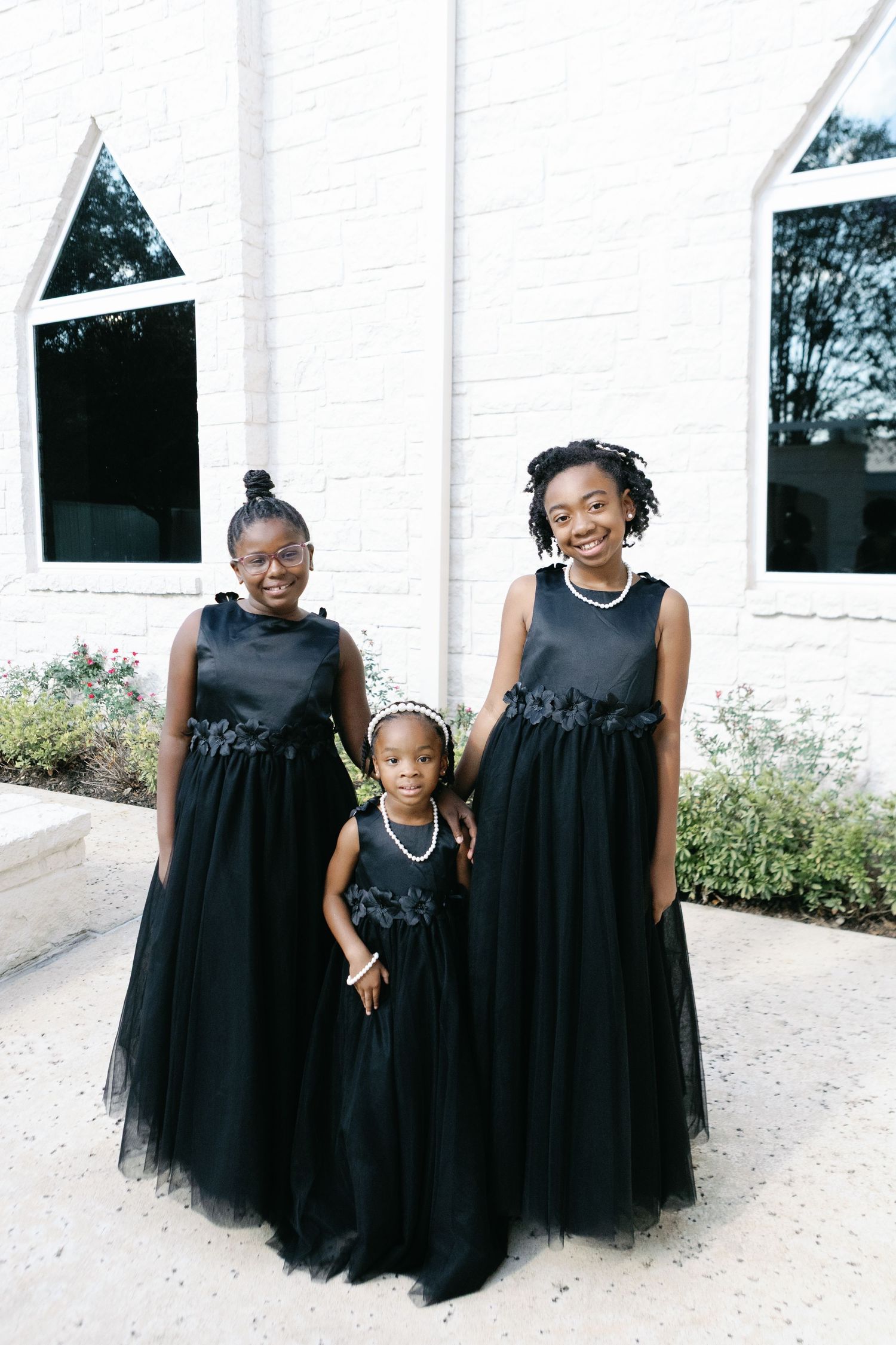 Young bridesmaids in matching black tulle dresses pose together against a white exterior wall.