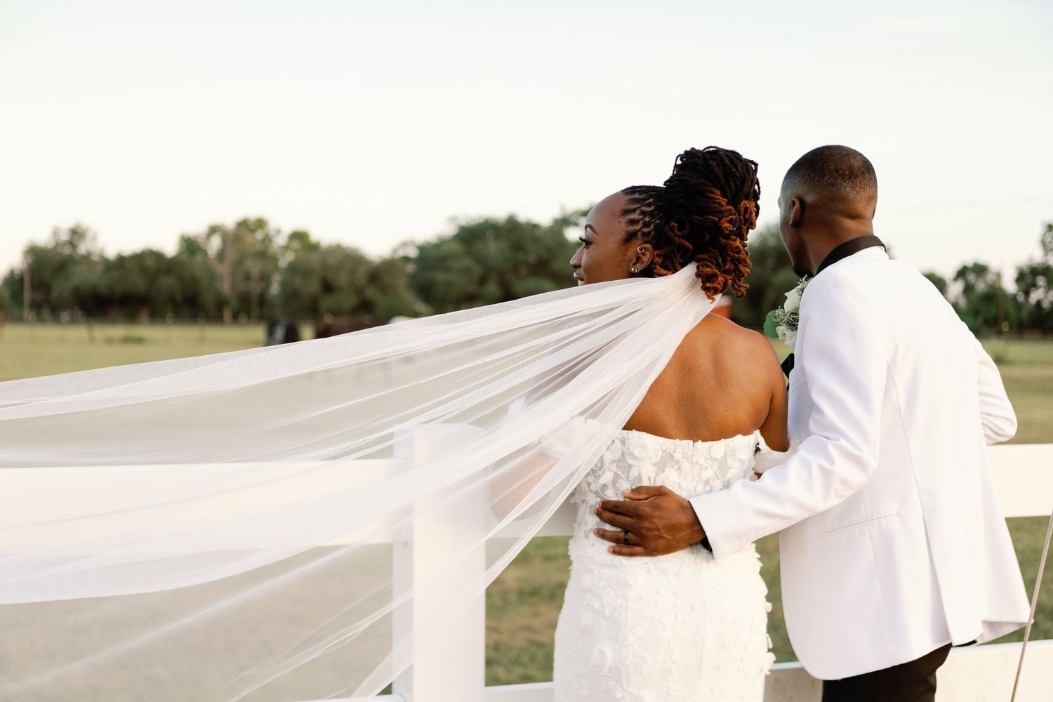 Flowing white wedding veil floats in the breeze as couple embraces against rustic outdoor setting.