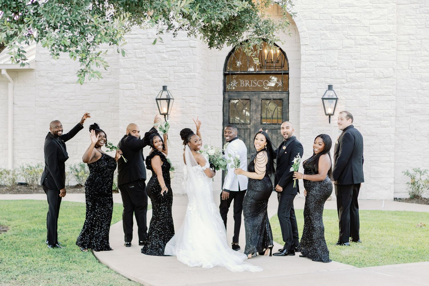 Wedding party dressed in black and white attire pose together by white stone wall with greenery overhead.