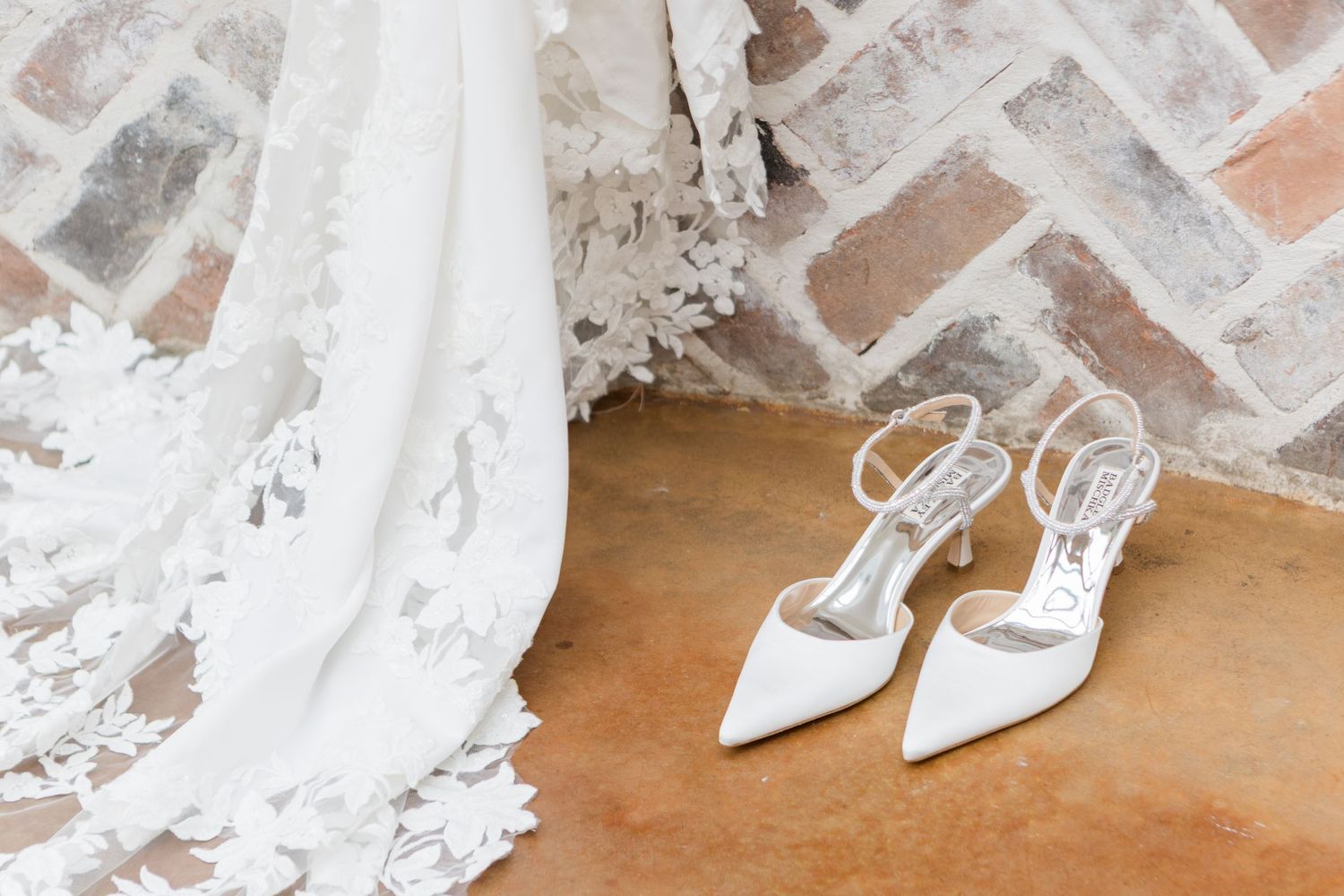 Close-up of white lace wedding shoes on a hardwood floor against exposed brick walls.