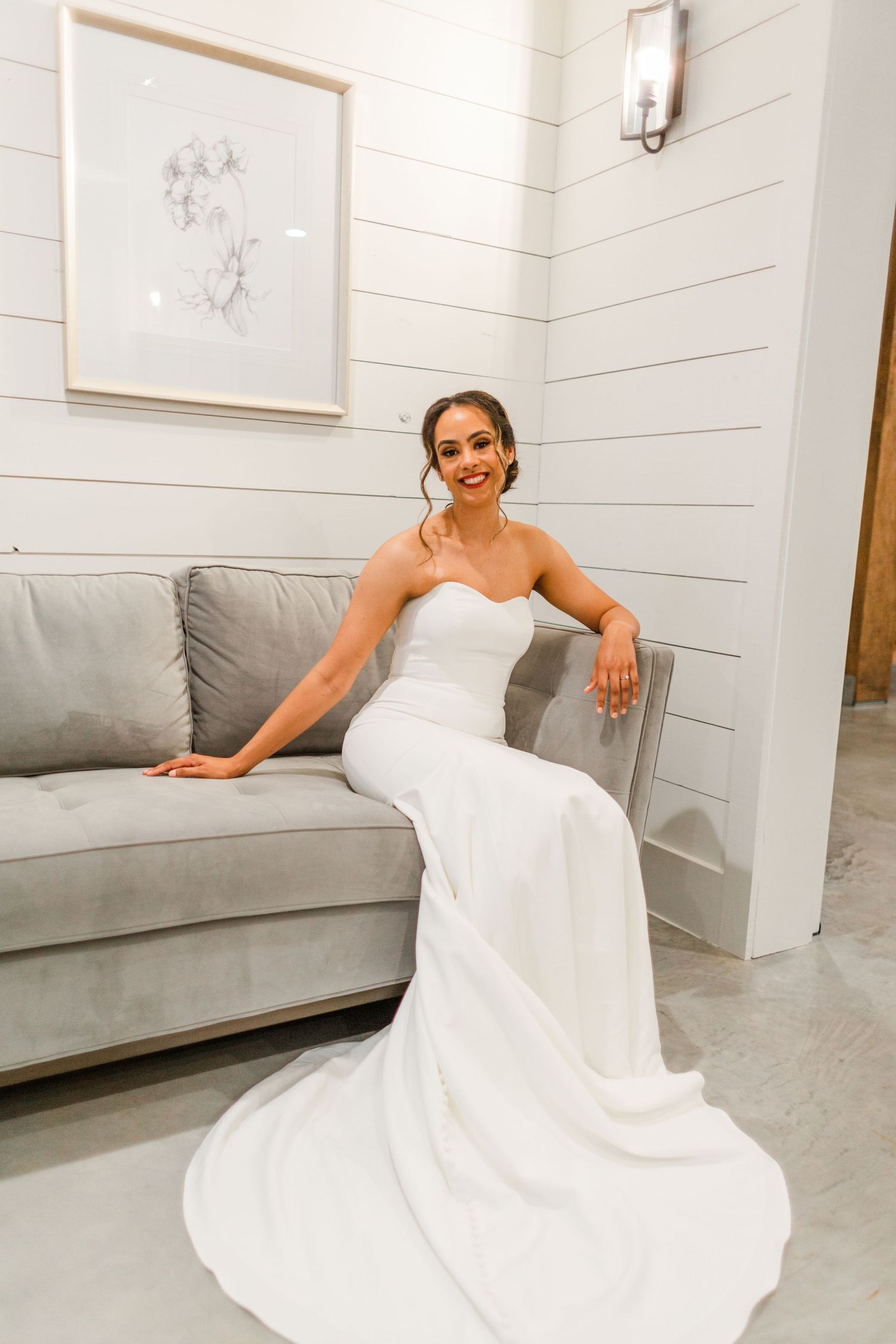 A bride in a white dress poses on a modern gray couch during a wedding photoshoot.