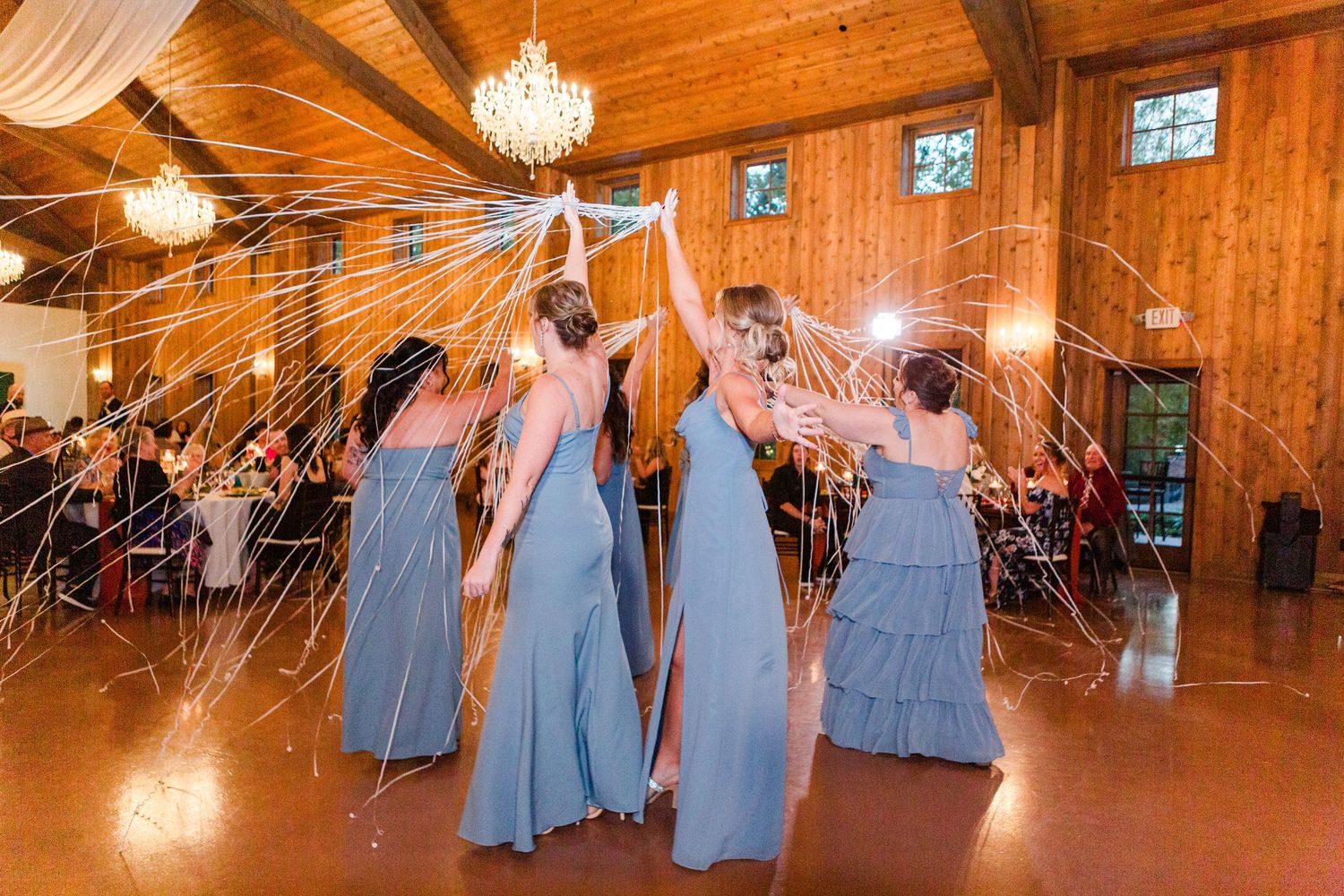 Bridesmaids in blue dresses dancing in a wooden reception hall with crystal chandeliers.