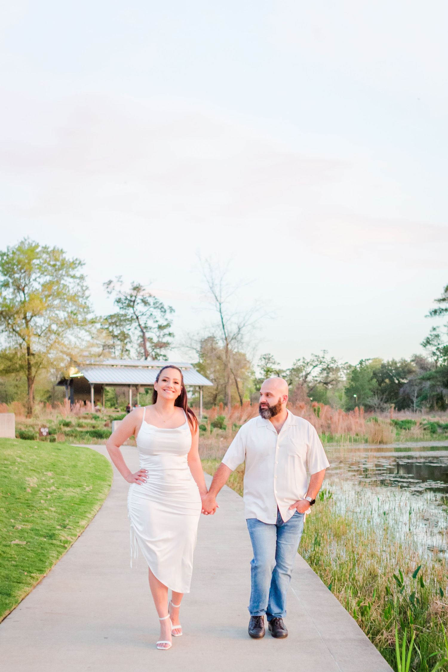Happy couple walking and laughing together along a lakeside path in white outfits.