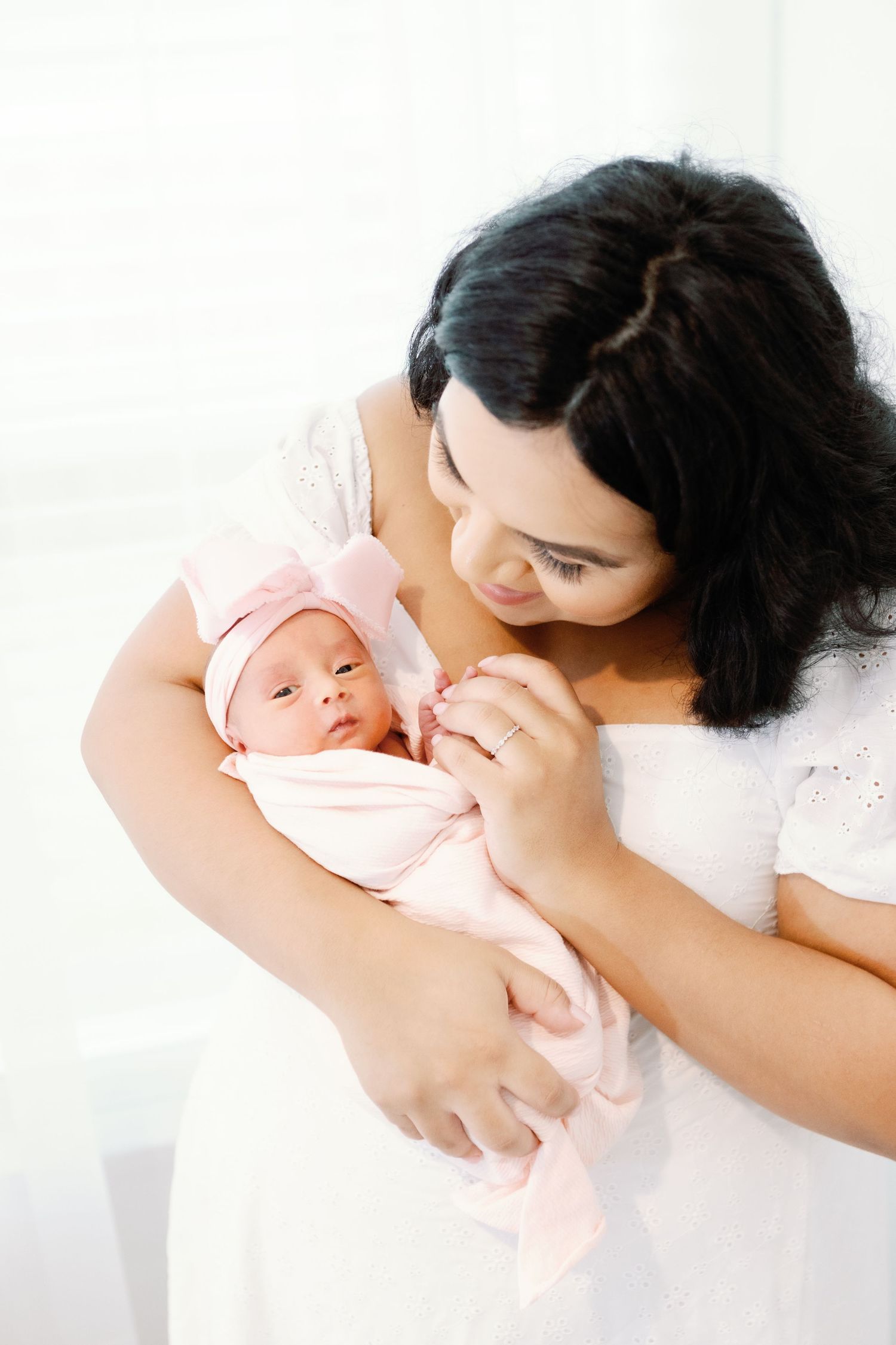 A parent and newborn baby share tender moments in soft pink tones while resting together on a white bedspread.