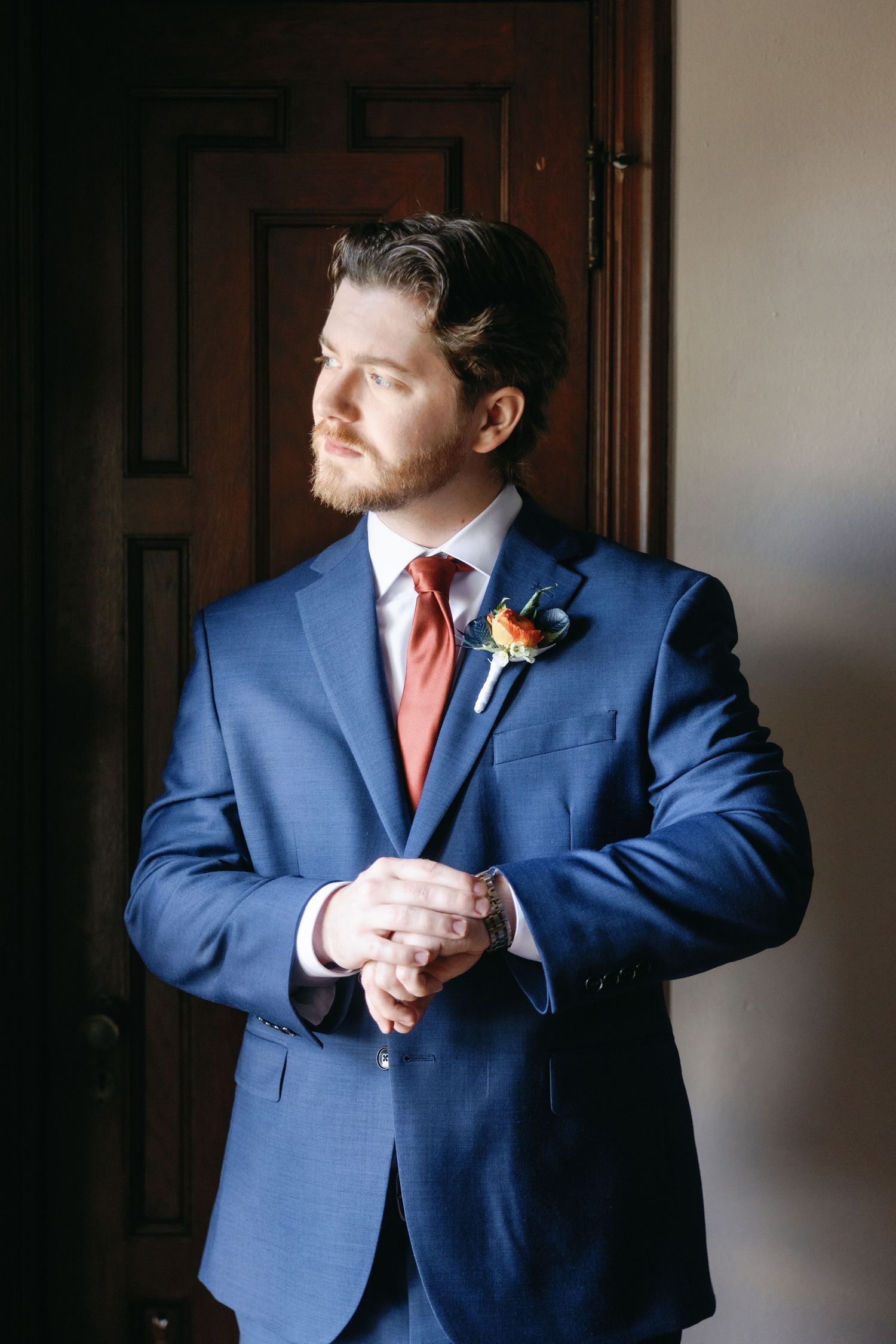 A groom in a navy blue suit and coral tie adjusts his cuffs while getting ready for the wedding ceremony.