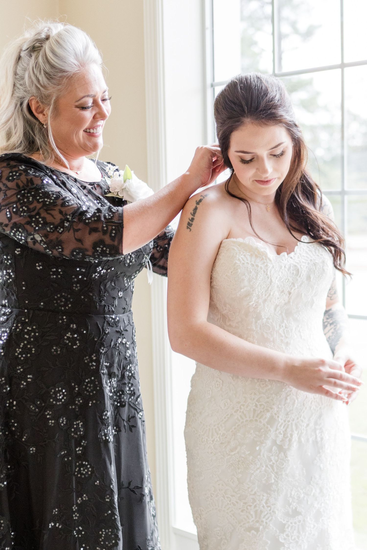 A woman in a black dress helps adjust details on a bride wearing a beautiful lace wedding gown.