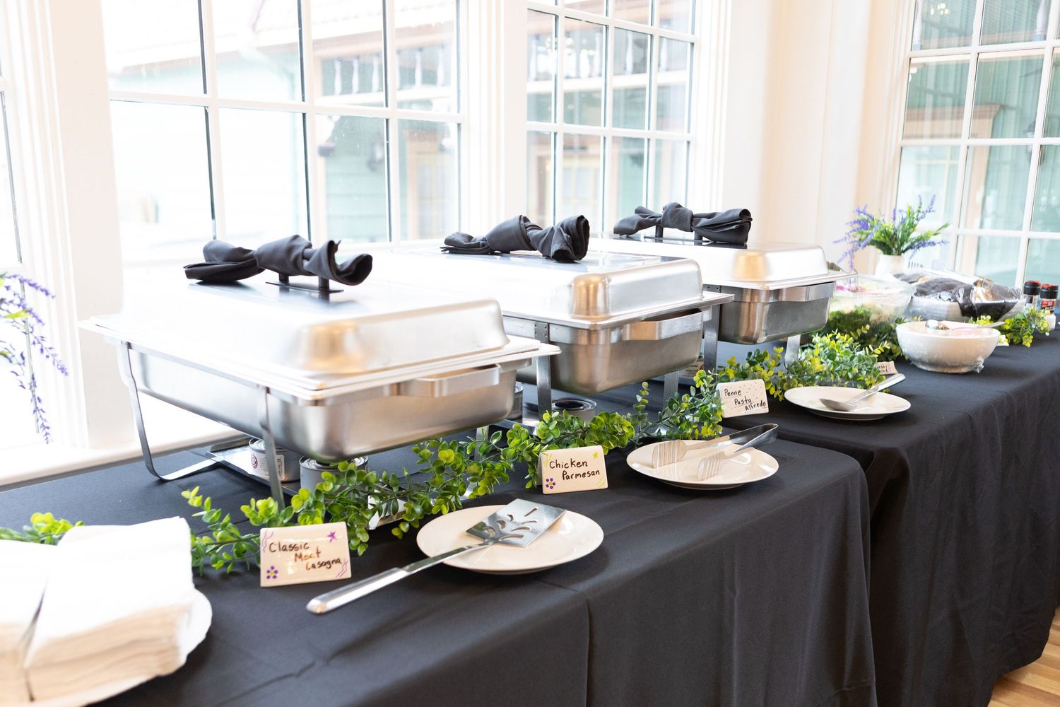 Wedding reception table setup with black linens and place settings near large windows.