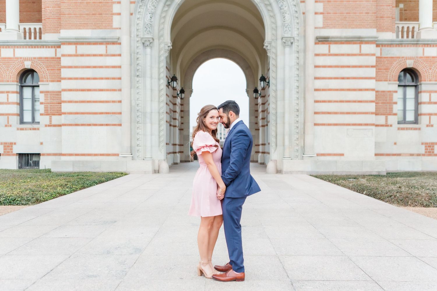 A couple embraces in front of ornate pink and white architecture with arched windows.