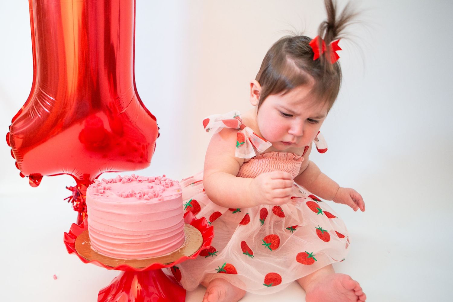 Baby reaching towards pink cake with red balloon and heart-print outfit.