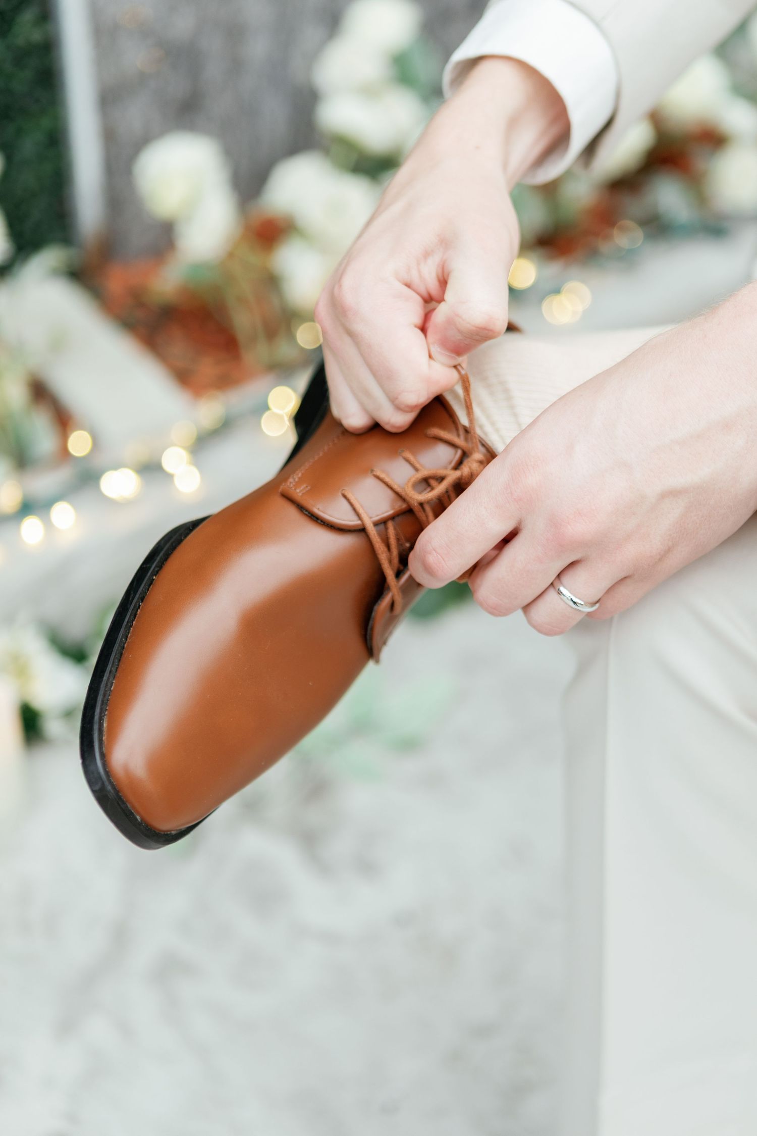 Close-up of hands tying brown leather dress shoe with wedding ring visible.
