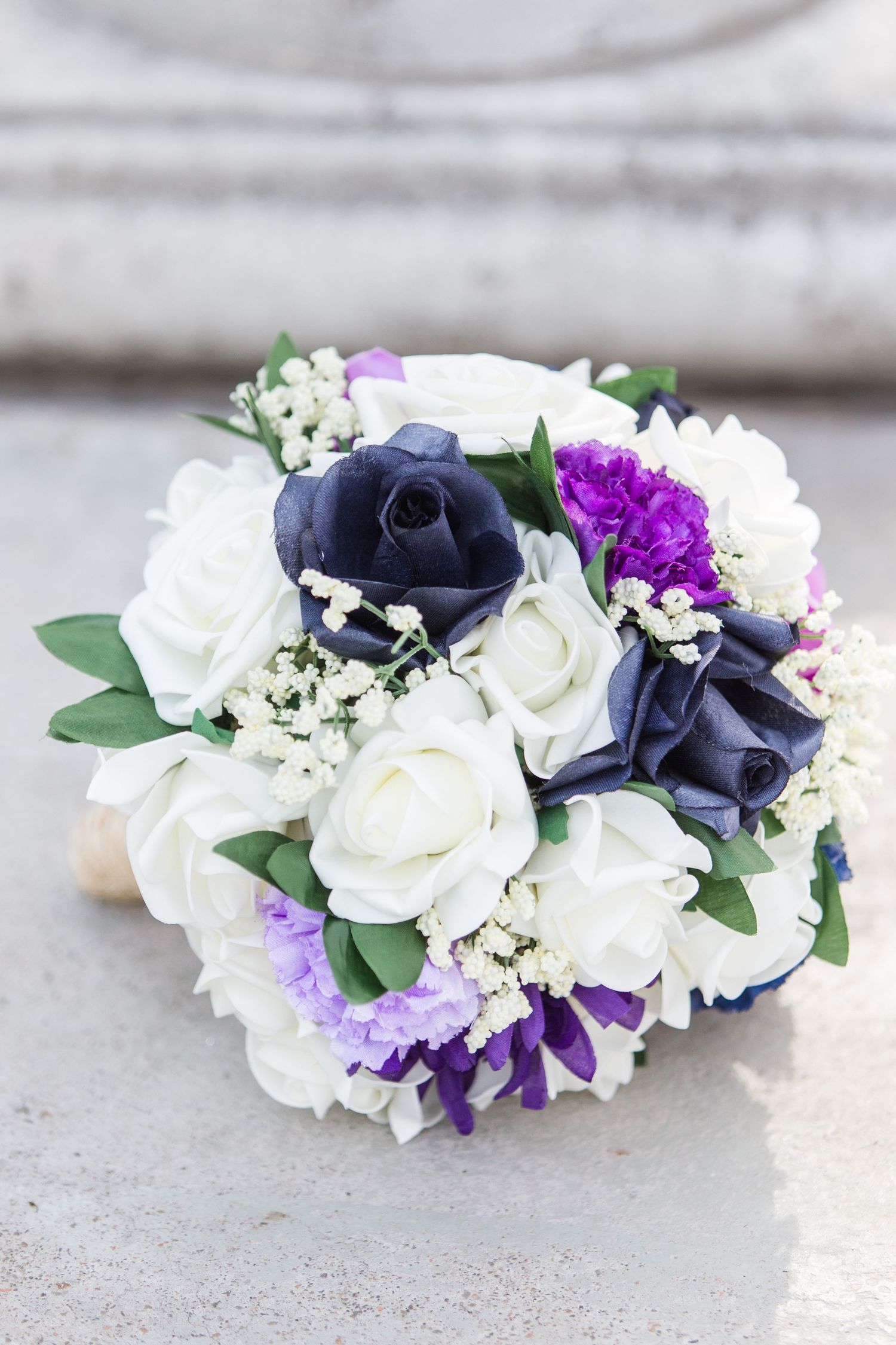 Floral crown made with white roses, navy blue flowers, and purple accents on a concrete surface.