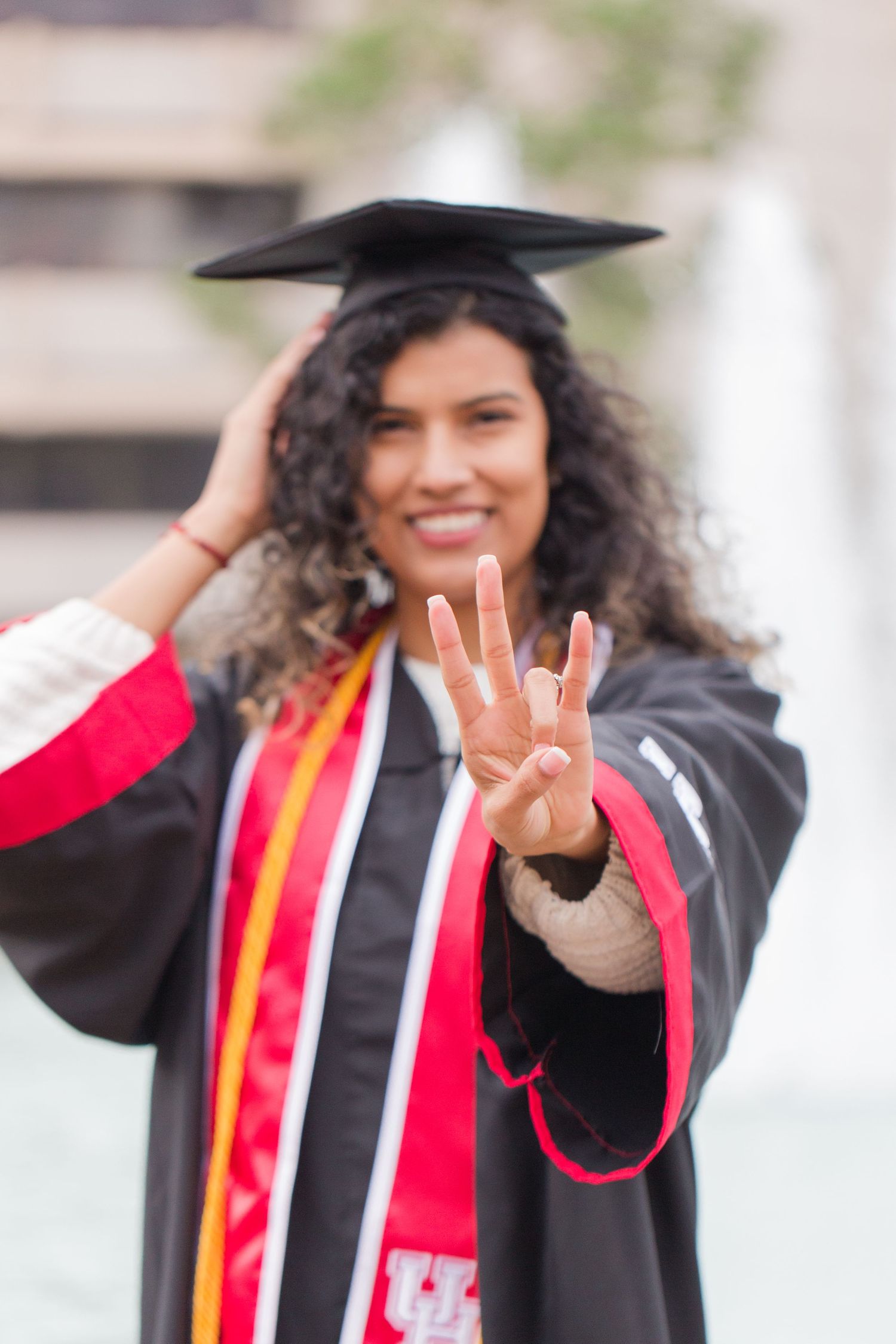 Graduate in black cap and gown with red stole poses excitedly during graduation photo session.
