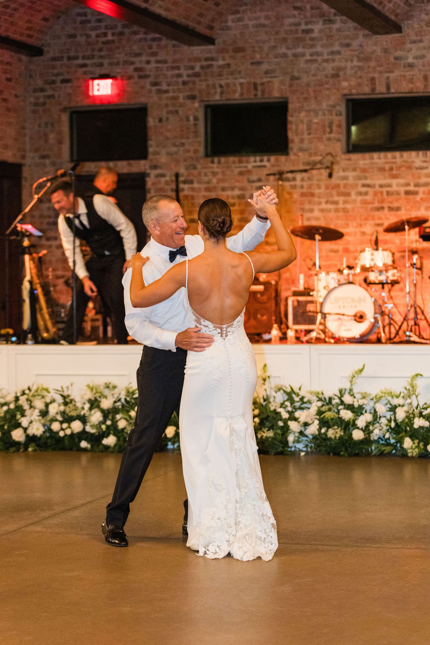 Couple dancing at a rustic brick-walled venue with floral decorations and band setup.