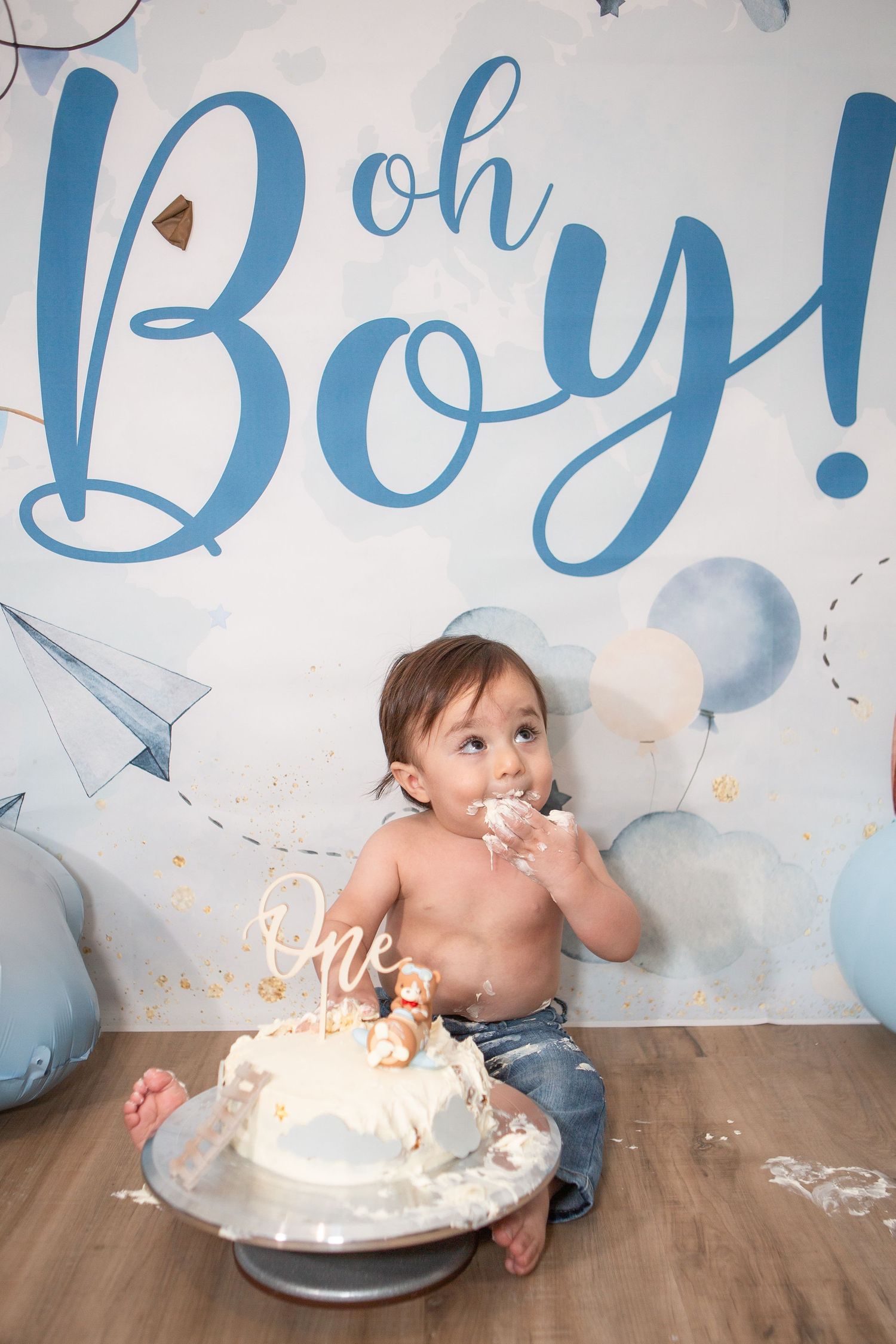 Baby sitting on wooden floor with demolished white birthday cake during cake smash session.