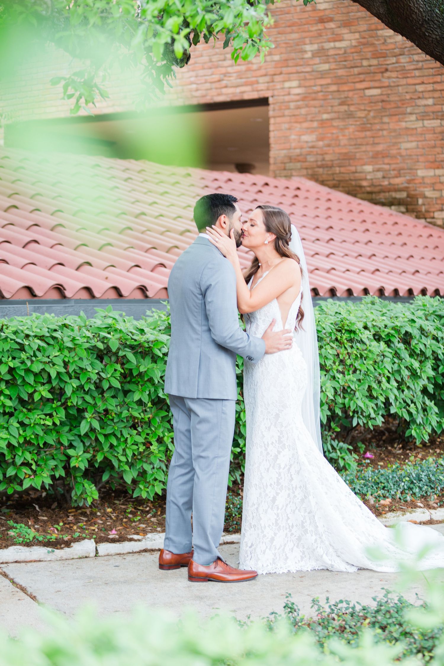 A couple sharing an intimate moment in front of a brick building with Spanish tile roof and lush greenery.