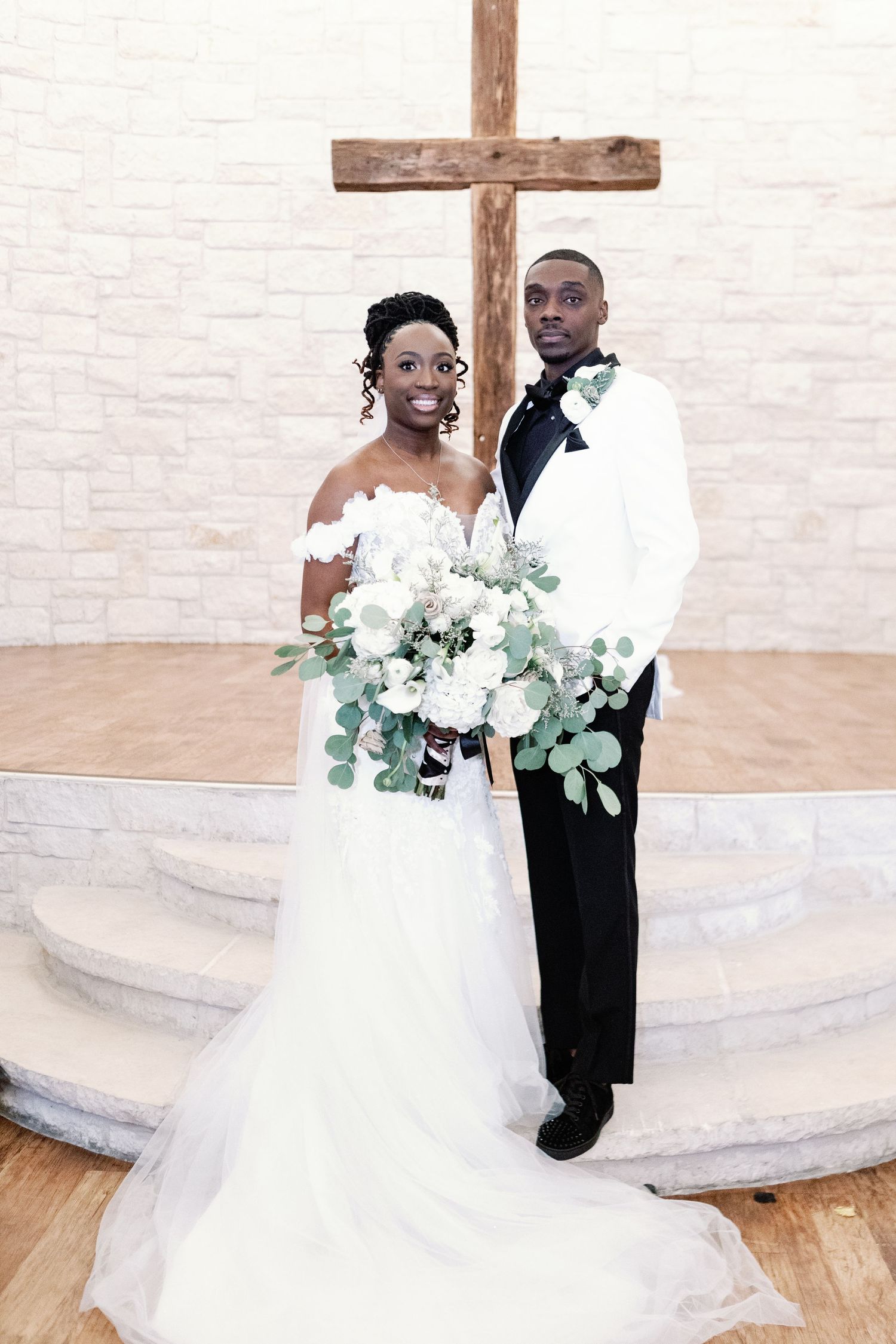 Wedding couple poses together in front of a wooden cross in a church with elegant white floral bouquet.