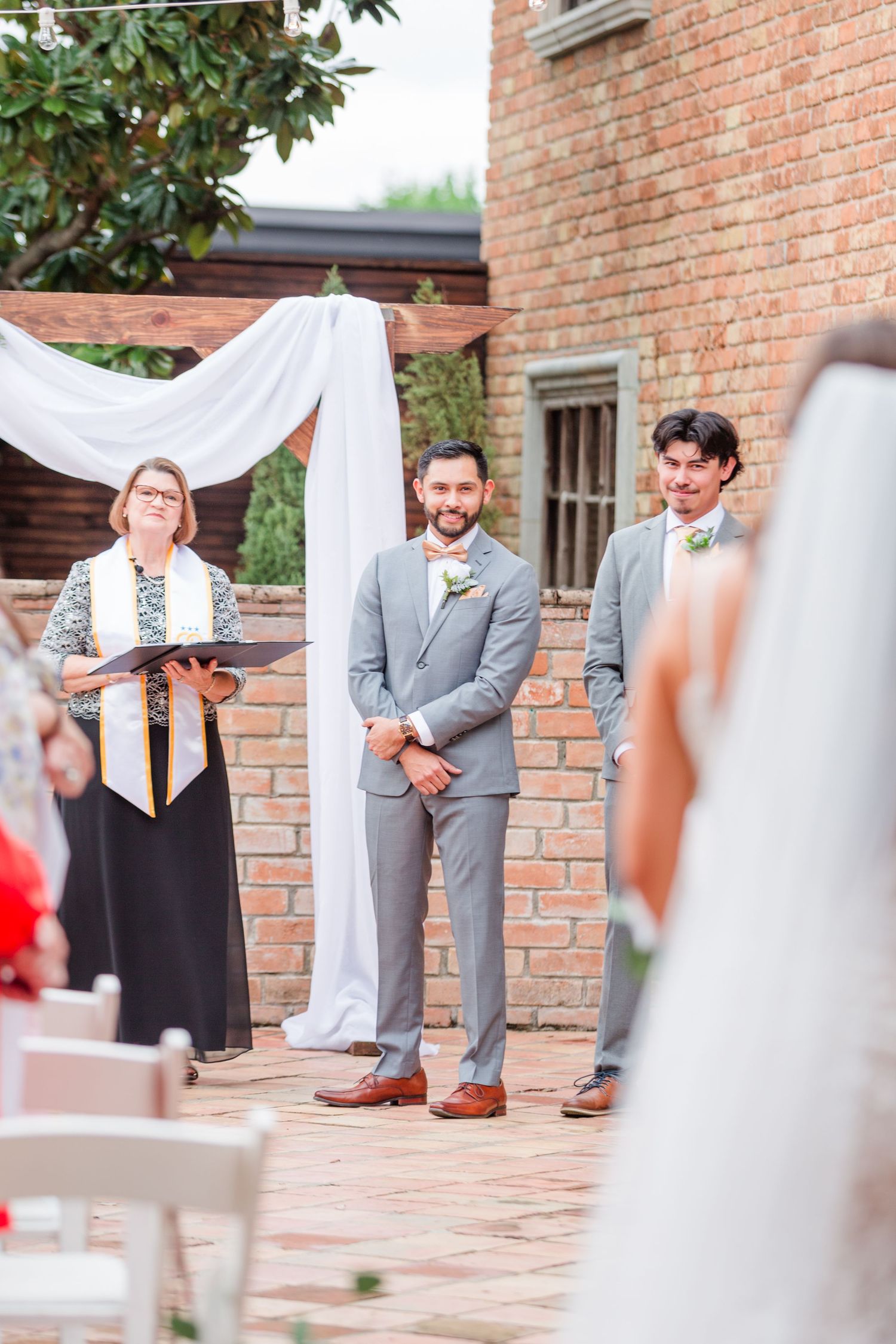 A wedding ceremony takes place outdoors by a brick wall with white drapery and floral decorations.