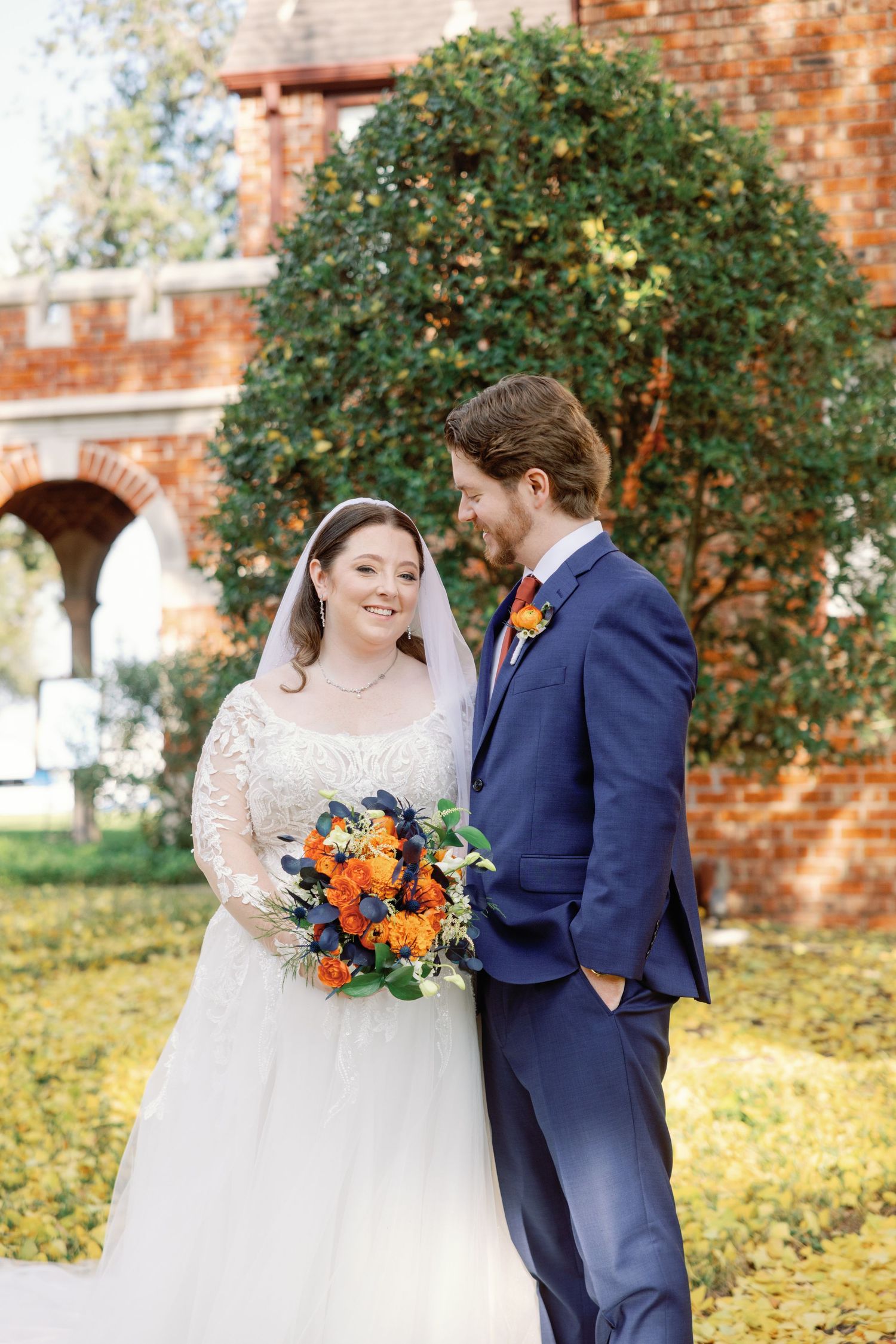 A couple shares a tender moment at their fall wedding with orange bouquet and navy suit against a brick backdrop.