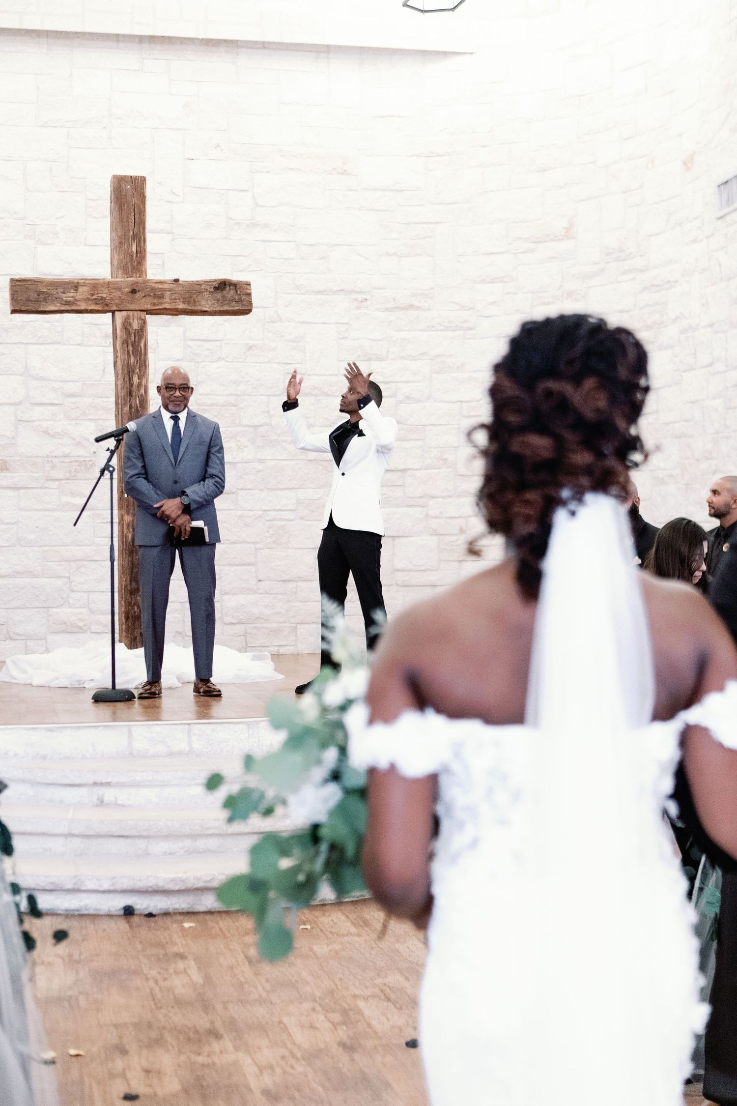 A wedding ceremony takes place in a minimalist white room with a wooden cross on the wall.