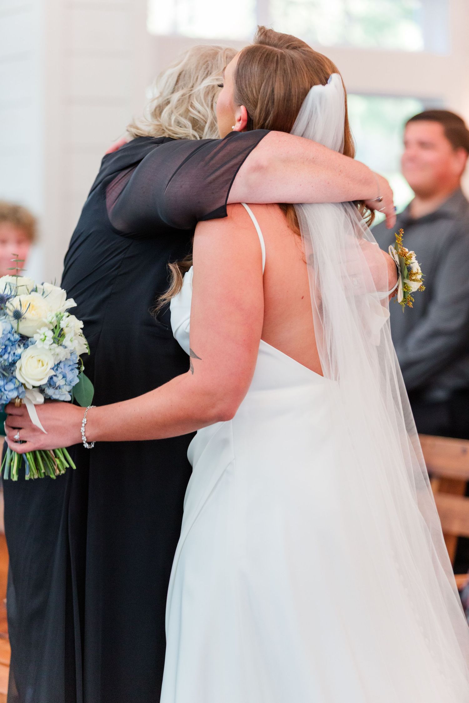 A bride in white dress and veil hugging someone while holding a blue and white bouquet.