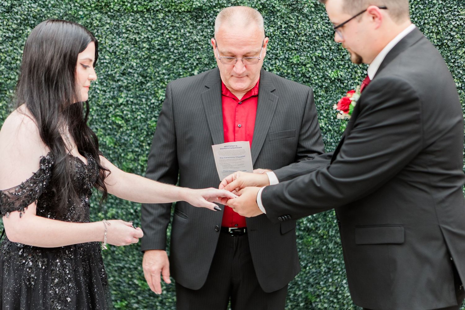 Wedding ring exchange ceremony being performed in front of lush green hedge backdrop.